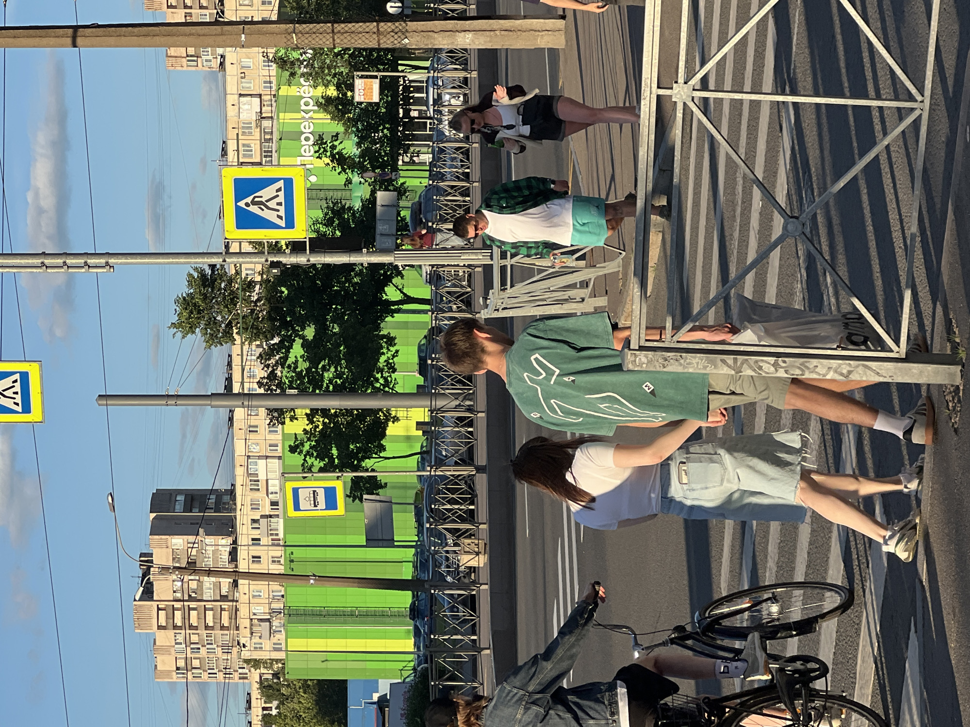 The image depicts a street scene in Sinyavino, Russia, bathed in the warm light of late afternoon or early evening. A group of people are casually gathered near a pedestrian crossing, suggesting a moment of pause or conversation.
The primary subjects are the individuals, their clothing indicating a relaxed, summery attire. One person wears a green plaid shirt and shorts, another a white t-shirt and denim shorts, and a third is seen in a green graphic t-shirt and light-colored shorts. A fourth individual, partially visible from the back, is wearing a denim jacket. The presence of a bicycle, with its wheel prominently featured in the foreground, hints at cycling as a mode of transport or a leisure activity.
The setting is urban, characterized by multi-story apartment buildings with many windows, typical of Russian residential areas. A prominent green-colored building is visible in the background, likely a commercial establishment or a modern renovation. Street signs are present, including a blue and yellow pedestrian crossing sign and another blue sign with a white icon of a bus or tram, indicating a public transportation stop. Electrical wires crisscross the sky, adding to the urban atmosphere. The ground is paved, with a crosswalk clearly marked, and shadows are cast across the scene by the low-angled sun and a metal fence, creating a play of light and dark.
The overall mood seems relaxed and unhurried, with the people appearing to be enjoying the pleasant weather. There is no clear indication of a specific event, but rather a snapshot of everyday life in a Russian town. The visible text includes Cyrillic lettering on the green building, which reads "Перекрёсток" (Perekrestok), meaning "Crossroads."