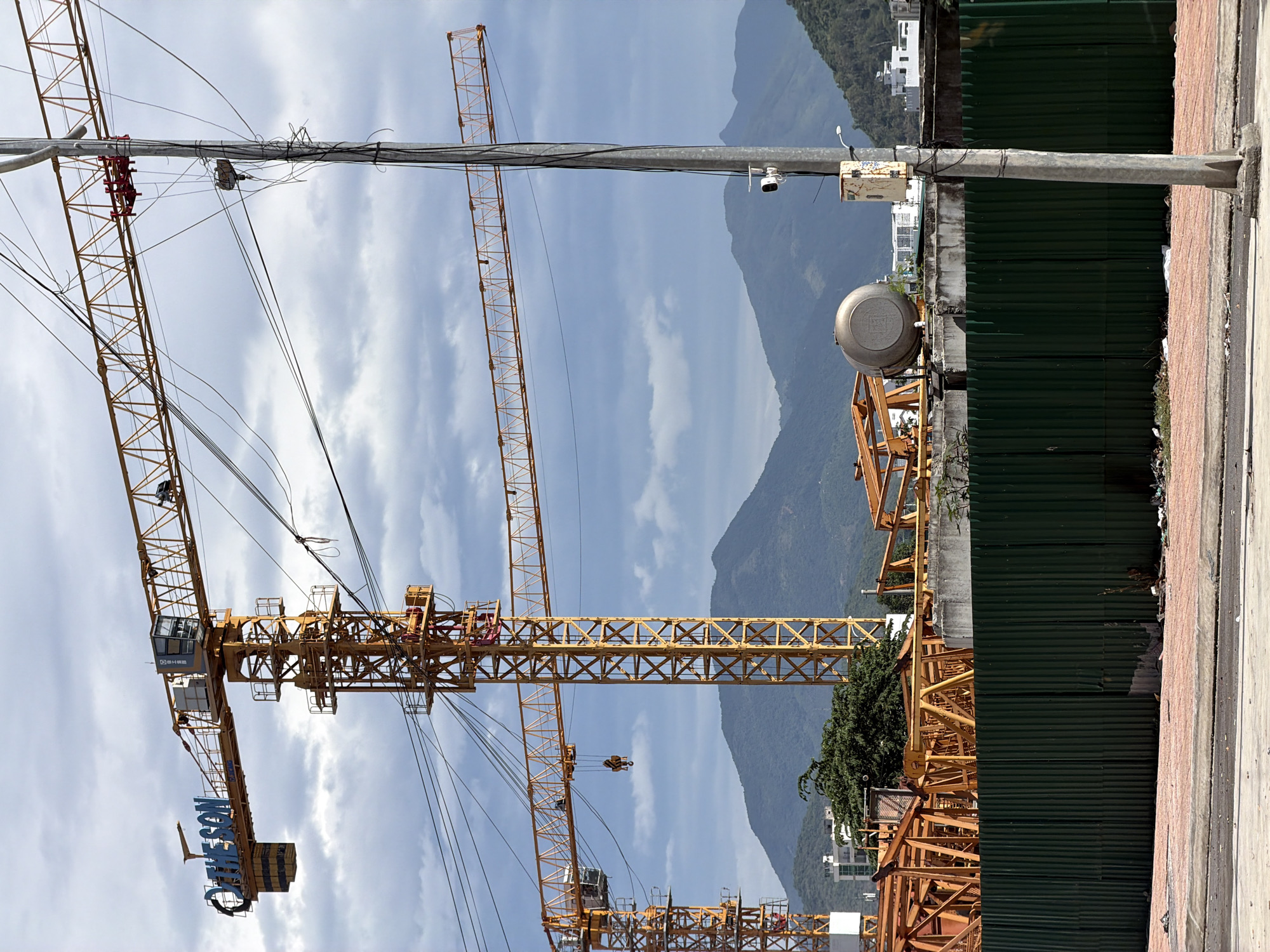 The image captures a bustling construction site in Nha Trang Ward, Vietnam, set against a backdrop of majestic mountains and a partly cloudy sky. Several large, yellow tower cranes are the primary subjects, indicating ongoing development. One prominent crane arm clearly displays the brand name "JAMESON" in blue letters.
In the foreground, a utility pole runs vertically, supporting a dense array of tangled black electrical wires and a small grey junction box. Two security cameras are mounted on this pole. Adjacent to the pole, a sidewalk composed of concrete and brick pavers lines a tall green corrugated metal fence, which encloses the construction area. Behind the fence, a large, grey, cylindrical tank-like structure is visible, along with parts of other construction elements and some green foliage.
The distant mountains rise grandly, covered in lush greenery, with a few buildings visible on their lower slopes. The scene is illuminated by clear daylight, suggesting a bright and clear day without any visible human activity.