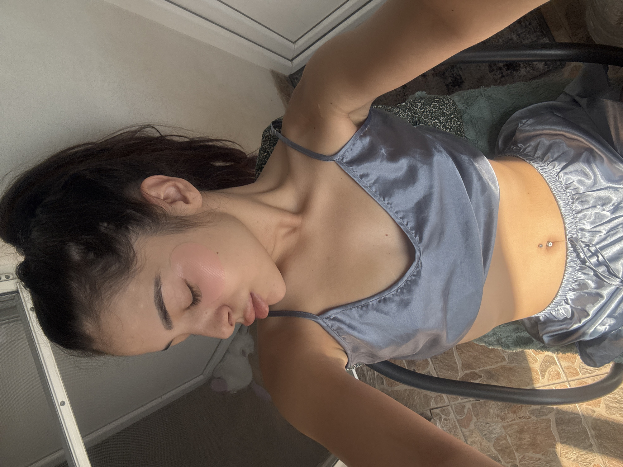 The image, taken in Plovdiv, Bulgaria, depicts a young woman relaxing indoors, captured from an overhead perspective. She is lying down, possibly on a chair or chaise, with her head turned to the side and eyes closed, suggesting a moment of rest or peaceful repose. She wears a light blue or slate-grey satin camisole with thin straps and matching satin shorts, characteristic of loungewear or sleepwear.
Notable details include light-colored, possibly translucent, skincare patches visible on her left cheekbone. Her dark hair is pulled back, revealing her ear. A small, shiny navel piercing is visible on her abdomen. Strong natural sunlight illuminates her body, creating clear highlights and shadows, indicating it is daytime. The background features a white-framed window or door on the upper left. Beneath her, parts of a dark metal chair frame are visible, along with a greenish patterned rug or blanket, resting on a tiled floor, possibly terracotta or a similar material. The scene conveys a relaxed and sunlit indoor environment. No text is visible in the image.
