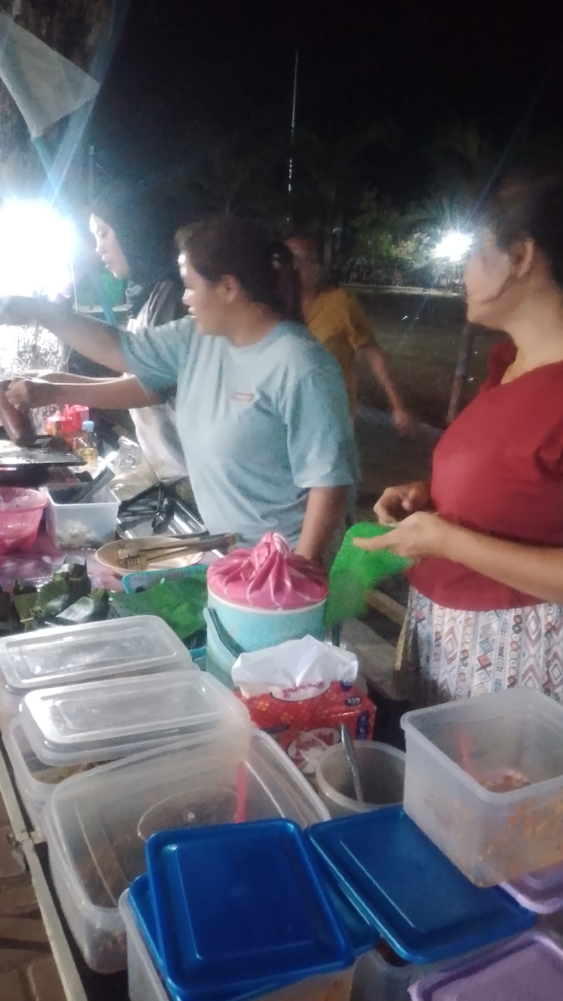 At a night market stall in Karanglangu, Indonesia, two women are engaged in food preparation and service. The woman on the left, wearing a light blue t-shirt, is attending to a cooking surface, likely a grill or griddle. In the background, another person in a yellow shirt is visible. To the right, a woman in a red shirt is interacting with a green plastic bag, possibly holding ingredients.
The stall is laden with various food containers, including clear plastic tubs of different sizes, some with blue and purple lids. There are also wrapped food items, possibly in banana leaves, and a pink-covered bucket. A tissue box with a red pattern is also present. The scene is illuminated by bright lights, suggesting it's a nighttime market. Palm trees are visible in the background, indicating an outdoor setting.