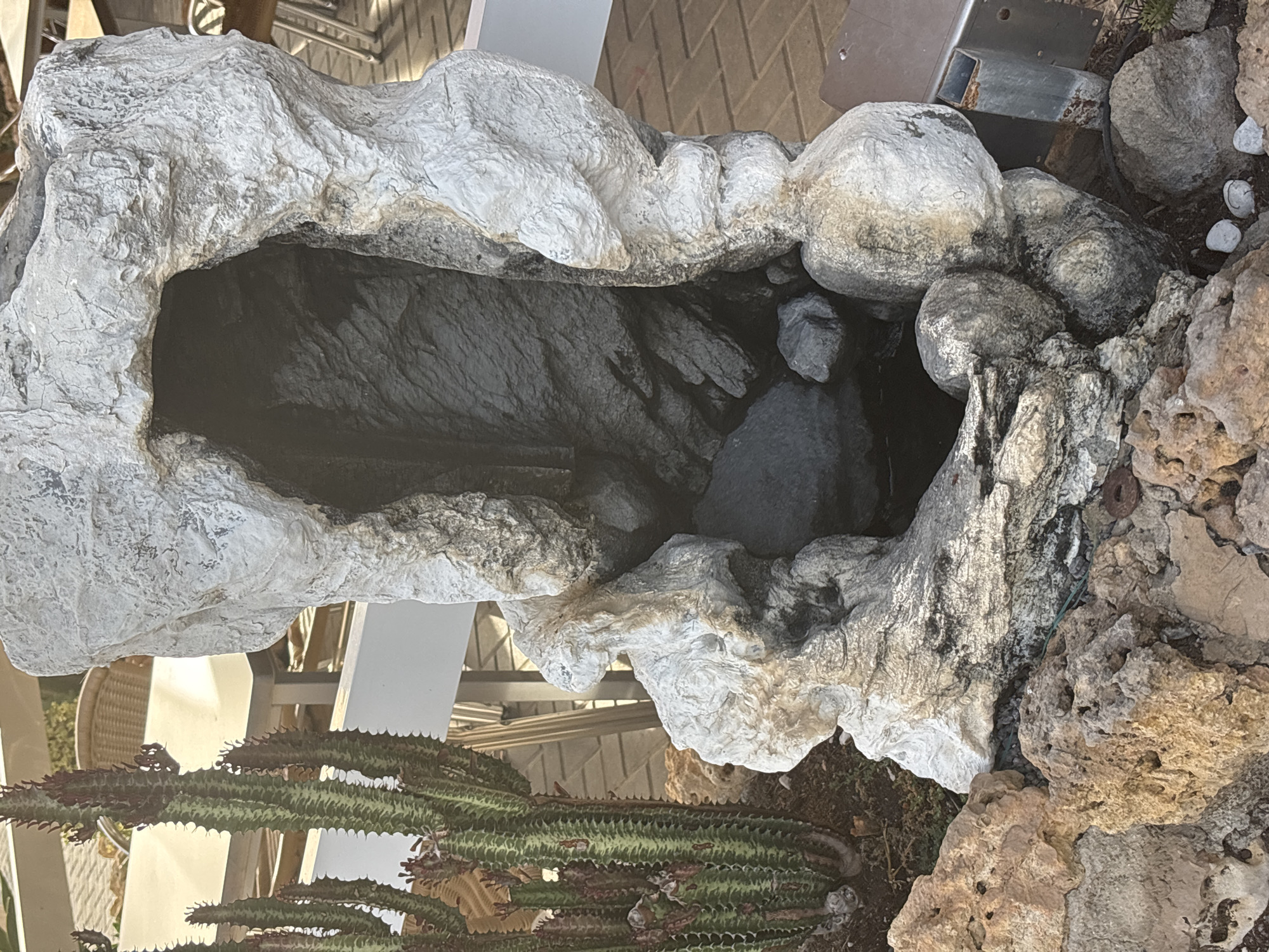 The image shows a close-up view of a man-made rock formation, resembling a cave or grotto, outdoors. The rock is textured and appears to be made of concrete or a similar material, painted a light gray with darker accents. Inside the opening of the rock formation, there is dark water, suggesting it might be part of a fountain or small pond. Surrounding the base of the rock formation are various natural rocks and pebbles of different sizes and colors, from light tan to dark gray. To the left of the rock formation, a tall, segmented cactus with green and reddish-brown hues is visible, planted in dark soil. In the background, out-of-focus elements of an outdoor seating area are present, including tables with light-colored tops and chairs with woven seats and metal legs. The ground surface appears to be paved with interlocking bricks. The lighting suggests it is daytime, with shadows indicating sunlight. There are no people present in the image. The overall scene depicts a decorative landscaping element in an outdoor public space, possibly a patio or garden area.