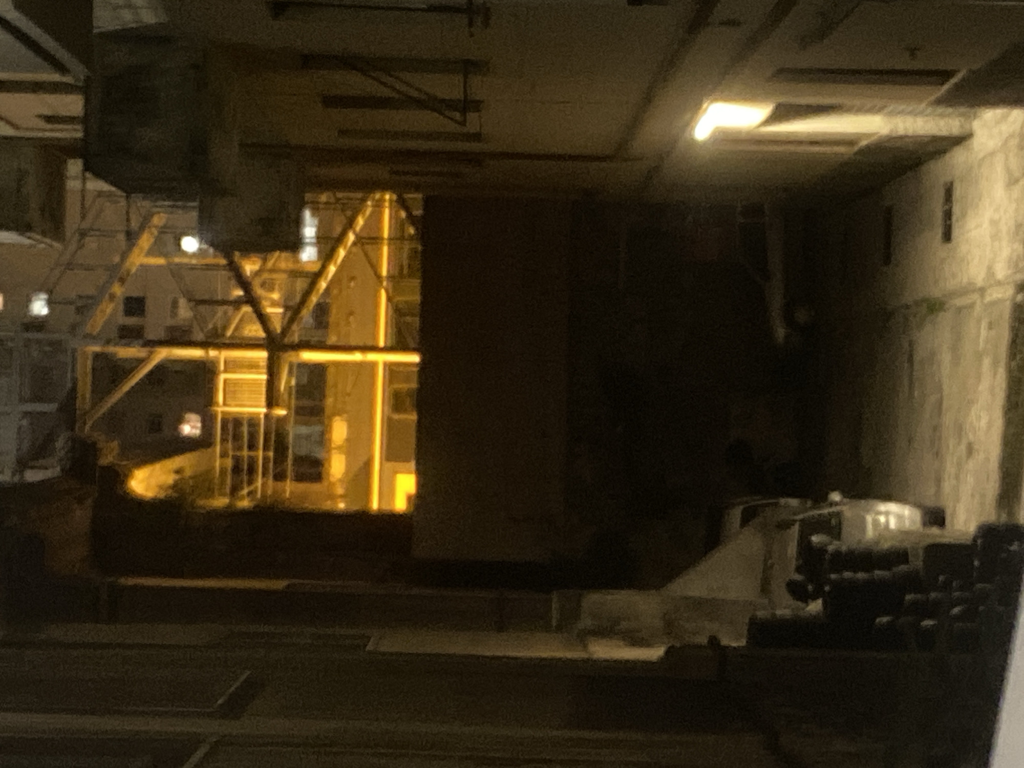 The image depicts a dimly lit alleyway in Genoa, Italy, at night. The perspective is from ground level looking down the alley. On the right side of the alley, a building with a lit window or lamp casts a bright yellow light onto the cobblestone ground. Further down the alley, another building or structure is visible, also illuminated with a bright yellow light, possibly from interior lighting or a construction site, as scaffolding is present. On the left side of the alley, a dark building wall dominates the foreground. In the middle ground of the alley, there are two vehicles parked. The one on the left appears to be a light-colored SUV or van, partially obscured by a dark, fence-like structure in the immediate foreground. The vehicle on the right is a darker-colored sedan, also parked in the alley. The overall impression is one of a somewhat desolate and utilitarian urban environment, likely in an older part of the city, characterized by its narrow streets and architecture.