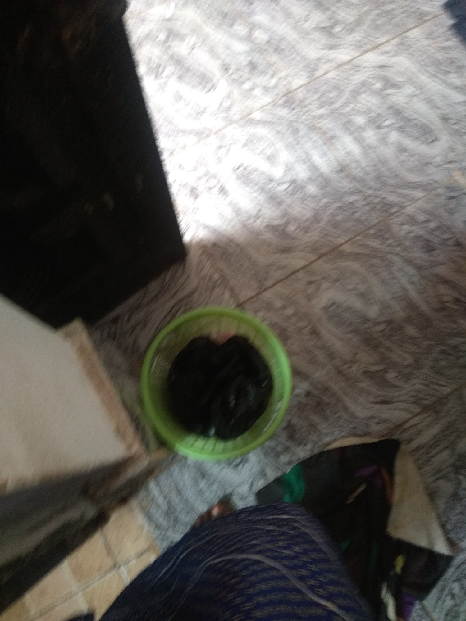A green plastic basket containing a dark, amorphous mass of what appears to be trash is situated on a patterned tile floor. To the left of the basket, a dark, rectangular object, possibly furniture or a cabinet, partially obscures the view. In the foreground, a person's legs are visible, clad in dark blue and purple patterned fabric. The overall scene suggests an indoor setting, likely a domestic space within the city of Jalingo, Nigeria, with the floor appearing to be tiled in a marble-like pattern. The lighting suggests an overhead light source, creating shadows and highlighting the textures of the floor.