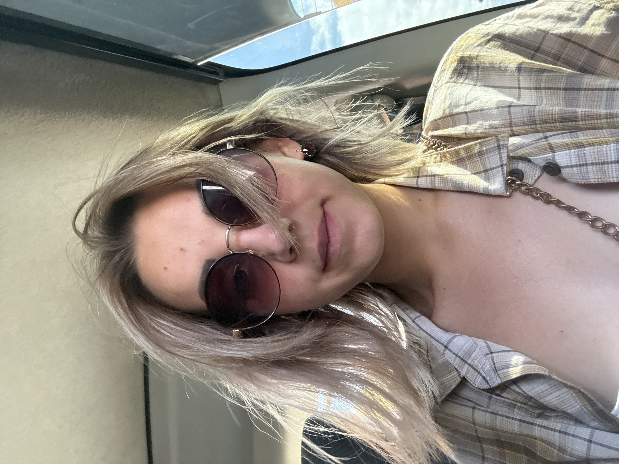 A young woman with shoulder-length blonde hair and sunglasses is taking a selfie inside a vehicle, likely a car, with a bright, cloudy sky visible through the window. She is wearing a light-colored plaid shirt with a chain necklace. The interior of the car appears to be the ceiling and a window frame. The location is the city of Yuzhno-Sakhalinsk, Russia.