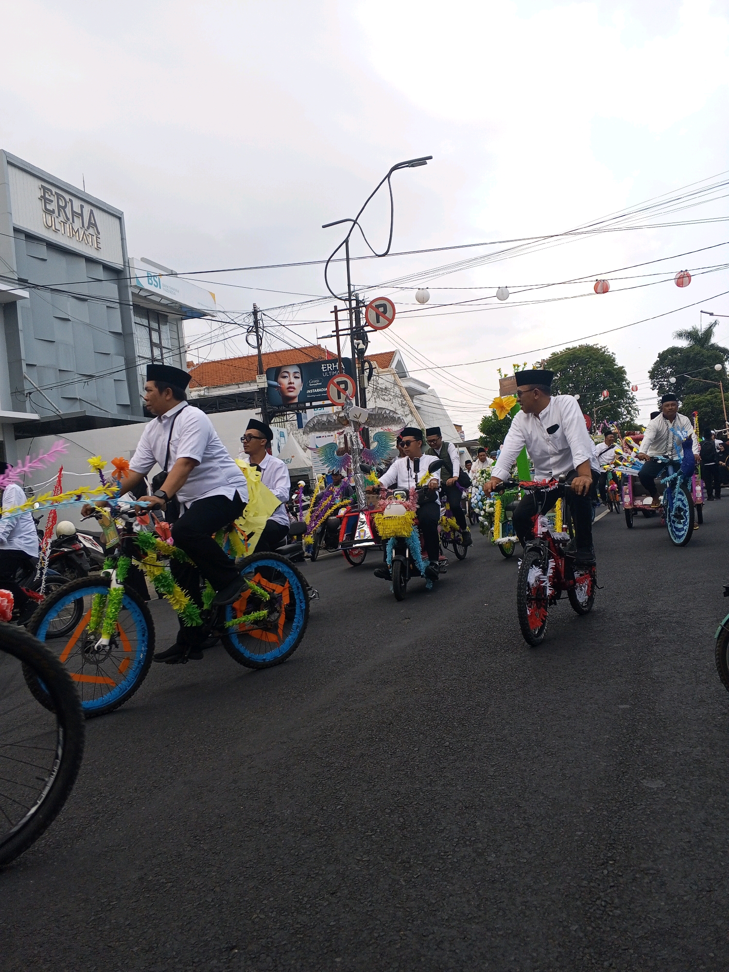 In Tamanbaru, Indonesia, a festive parade is underway on a cloudy day. A group of men, dressed in white shirts and black caps, ride decorated bicycles. The bicycles are adorned with colorful streamers, flowers, and other festive decorations. The men appear to be participating in a cultural event or celebration. The street is paved, and there are buildings and trees visible in the background. A prominent building with the signage "ERHA ULTIMATE" suggests a commercial area. The sky is overcast, and the overall atmosphere is one of community and celebration.