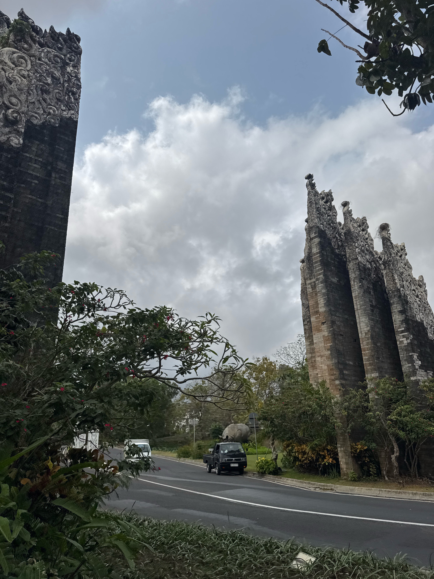 The image depicts a street scene in Kuta Selatan, Indonesia, featuring two prominent Balinese stone carvings on either side of a paved road. The carving on the left is a tall, dark brick structure with intricate stone ornamentation at the top. The carving on the right is also a stone structure, appearing to be comprised of multiple pointed spires and adorned with detailed carvings.
The road is asphalt with white lane markings and a gray curb. A black utility vehicle is parked on the right side of the road, and a white van is driving away from the camera on the left side of the road.
Green foliage and trees are visible on both sides of the road, with some red flowers blooming on the trees in the foreground on the left. The sky is overcast with a mix of gray clouds and some lighter patches. In the background, a large, gray, rhinoceros-shaped statue is visible on the left side of the road, behind some foliage. The overall environment suggests a tropical or subtropical location.
