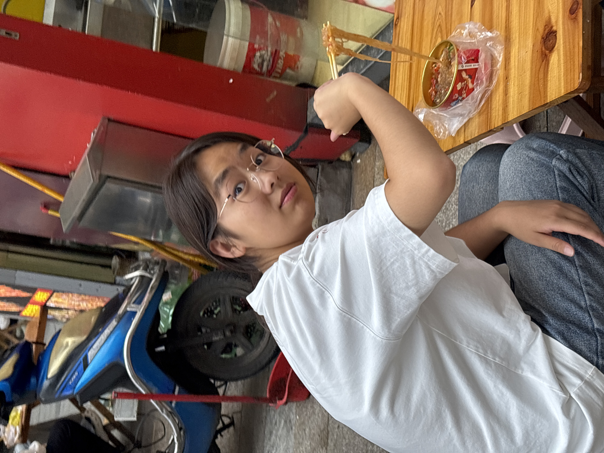 A young East Asian person wearing a white t-shirt and grey jeans is seated at a wooden table outside a food stall in Jiangjin, China. They are holding chopsticks with noodles, about to eat from a red can placed in a plastic bag on the table. In the background, a blue and silver scooter is parked next to a red food stall structure. The ground is paved with grey stones. The person is looking at the camera with a neutral expression.