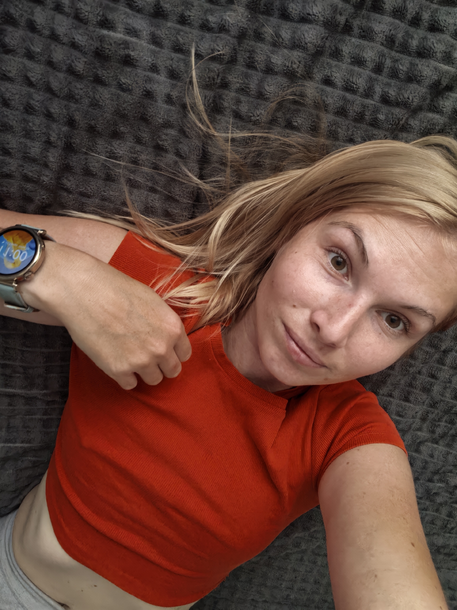 A young woman with blonde hair and light brown eyes is lying on a textured grey blanket, taking a selfie from a high angle. She is wearing an orange t-shirt and grey shorts, and a smartwatch on her left wrist displays the time "11:00". The background is entirely the grey blanket. No elements in the image suggest a location in Sterlitamak, Russia.