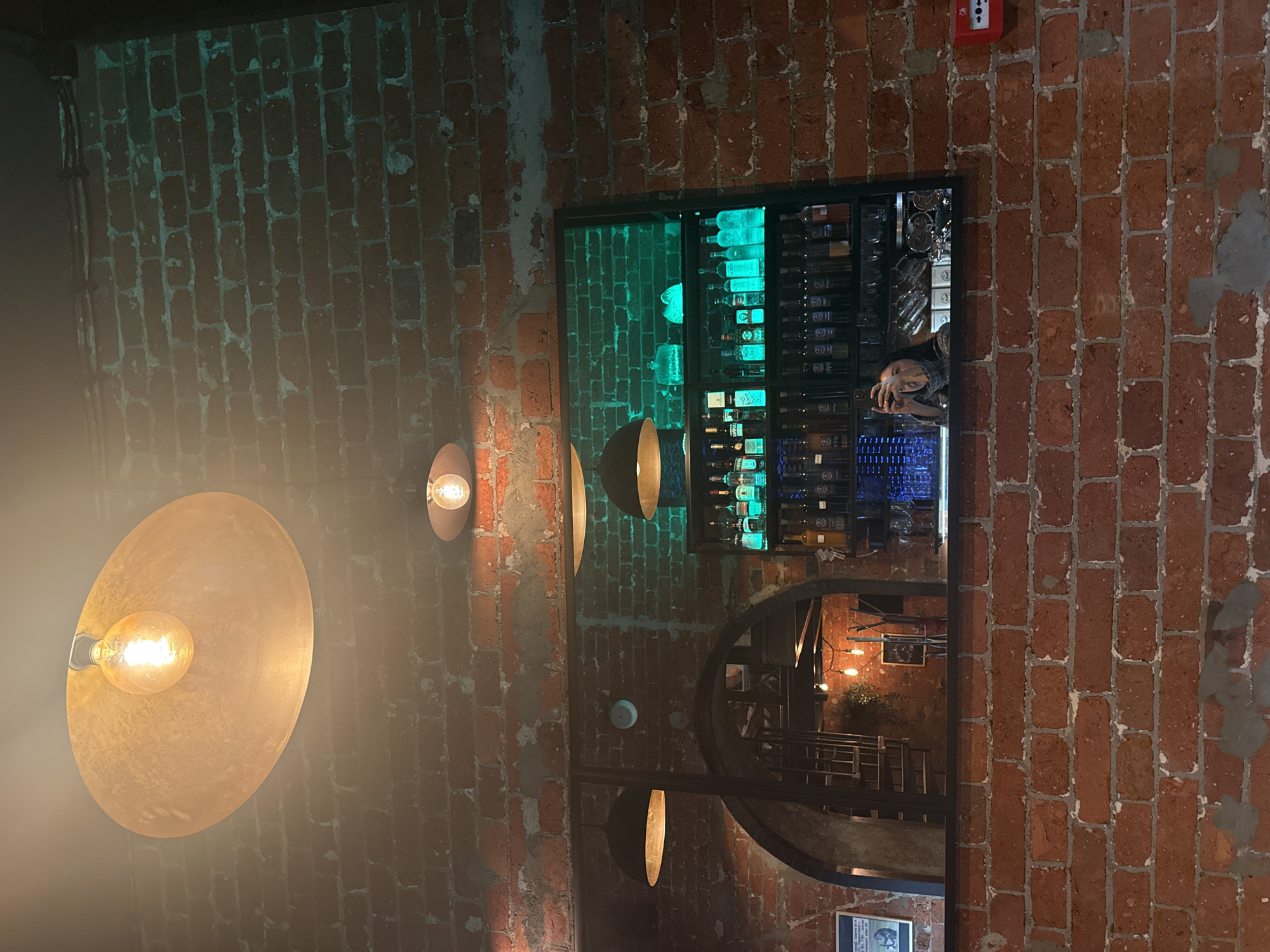 A vertical, medium-shot depicts a brick wall interior with two exposed-bulb pendant lights and a framed mirror reflecting a bar. The brick wall is red and has sections of light-colored mortar. The leftmost pendant light is a large, shallow, brass-colored disc, with a bright, glowing bulb visible in the center. To the right of the large pendant, there is a smaller, mushroom-shaped pendant light. The mirror is rectangular and framed in black. The mirror reflects a bar setup with bottles of liquor on a shelf that is illuminated with a green light. A person is reflected in the mirror, taking a photo of the space, as well as a stairway that leads to a lower floor with additional lighting. There is also a red fire alarm box on the wall, near the top right corner. A white sign can be seen reflected on the bottom edge of the frame.
Without additional information, it is not possible to confirm the location in the city of Moscow, Russia.