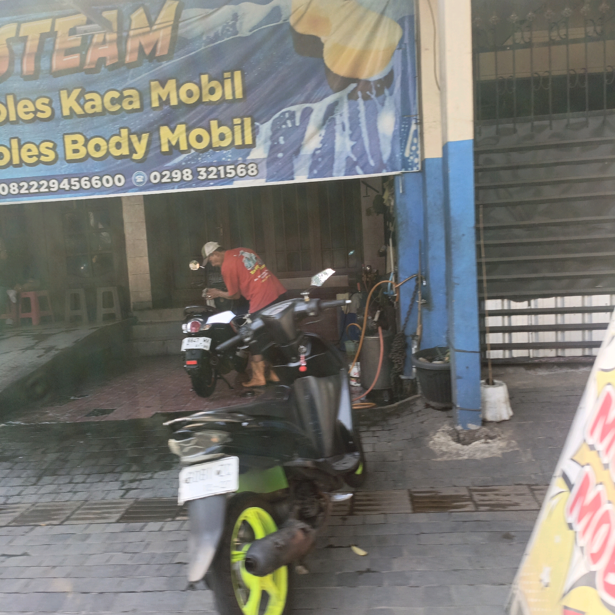 The image depicts a car and motorcycle wash service, likely located in Salatiga, Indonesia, based on the text on the banner. The banner, displaying "STEAM" in large yellow letters and services like "bles Kaca Mobil" (glass car wash) and "bles Body Mobil" (body car wash), hangs above the service area. A man in a red t-shirt and a white cap is seen washing a motorcycle parked in front of a building with a blue pillar and metal gates. Another motorcycle, dark colored with a white license plate, is parked in the foreground. The ground appears wet, suggesting active use of the washing facilities. The overall scene conveys a typical street-side service business.