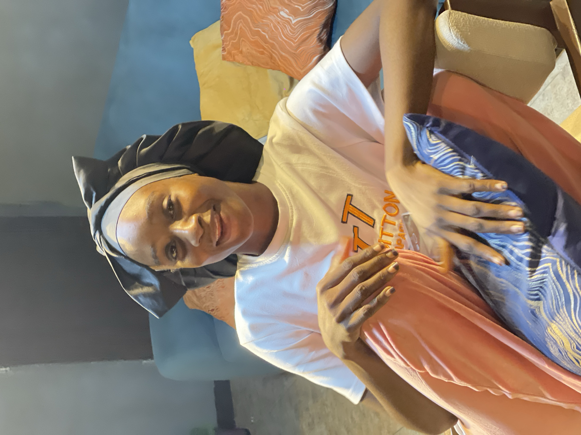 A young Black woman with a satin bonnet is sitting in an armchair, looking at the camera with a smile. She is wearing a white t-shirt with an orange graphic and peach-colored pants. Her hands are resting on a blue and gold patterned pillow. In the background, a teal sofa with yellow and orange accent pillows is visible. The scene appears to be indoors, possibly in a residential setting in Abuja, Nigeria. The lighting is soft, creating a warm and relaxed atmosphere.