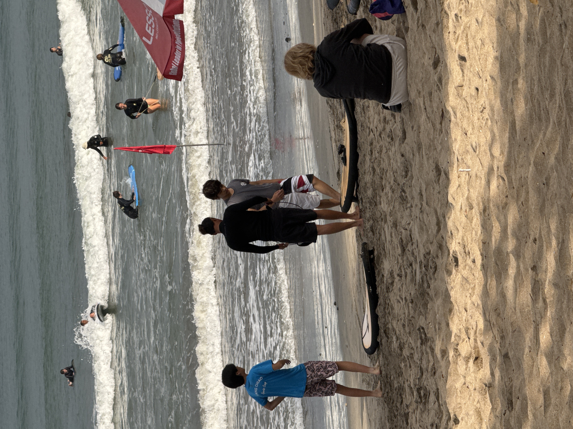 The image depicts a beach scene in Kuta, Indonesia, with several individuals engaged in or preparing for surfing. In the water, there are multiple people on surfboards, some riding waves and others waiting. Closer to the shore, three young males are standing on the sand. One, wearing a blue t-shirt with text and patterned shorts, faces away from the camera. Another, in a black wetsuit and black shorts, also faces away. The third, wearing a grey t-shirt and striped shorts, is seen in profile. A woman with blonde hair is seated on the sand, facing away from the camera, wearing a grey hoodie and light-colored pants. A red flag is planted in the sand, and a red and white canopy, possibly from a beach vendor or surf school, is visible in the foreground. Two surfboards lie on the sand near the seated woman. The ocean exhibits moderate waves, and the sky appears overcast.