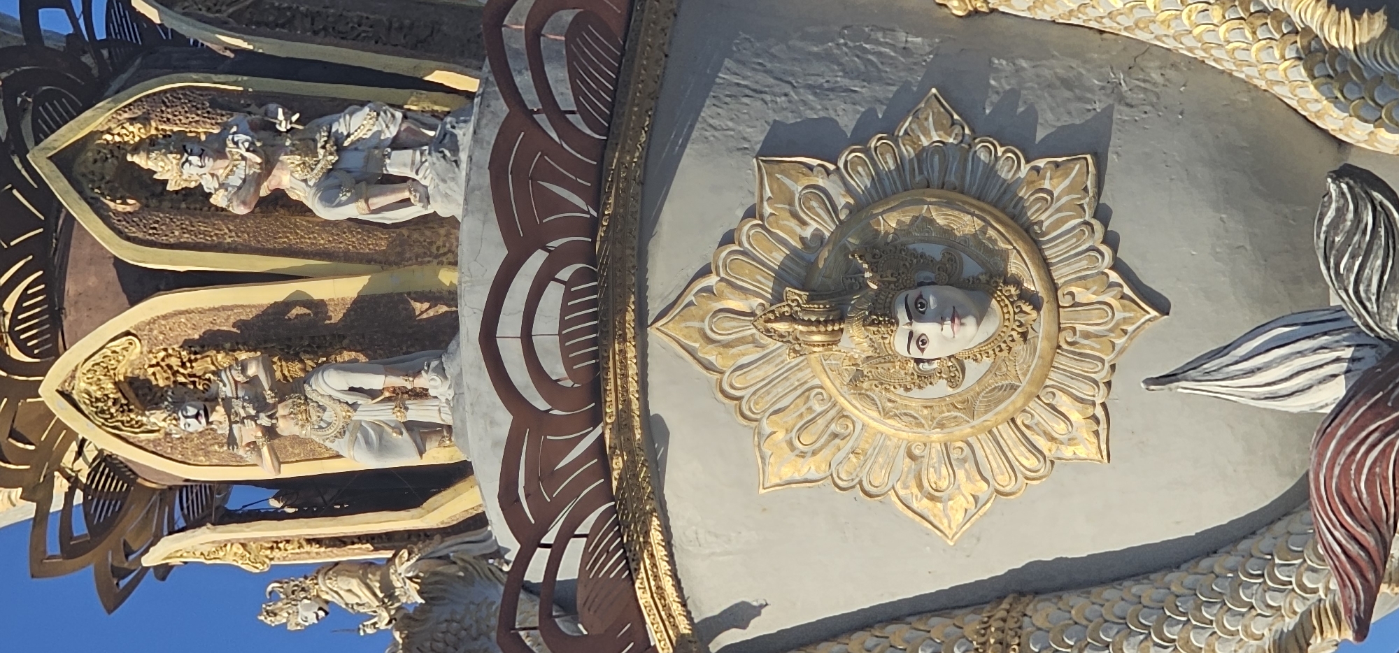 The image shows a close-up of a temple in Denpasar, Indonesia. The architecture is elaborate, featuring carved figures, floral designs, and a representation of a face in the center of a decorative sun or flower. The color palette is predominantly gold, white, and shades of grey, with accents of red-brown. The lighting suggests it is daytime, and the angle is looking upwards.