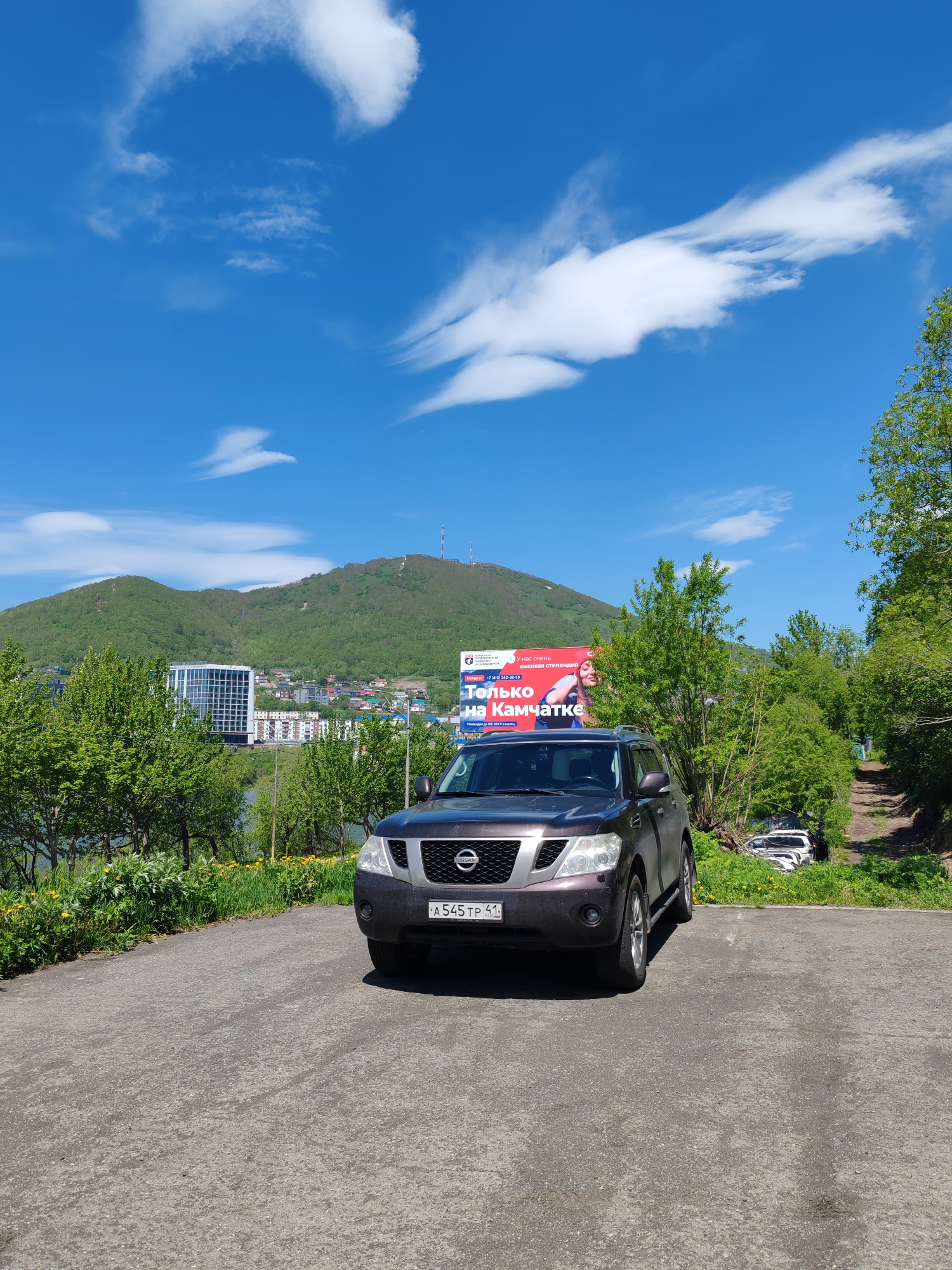 An eye-level, front-facing medium shot captures a dark brown Nissan Armada SUV parked on an asphalt surface. The SUV is positioned centrally in the frame, with its front end facing the camera. Behind the vehicle, a vibrant green hillside dominates the background, dotted with scattered trees and buildings. A large billboard with a picture of a woman and Russian text is visible on the right side of the frame. The sky is a bright blue with scattered white clouds. To the right of the SUV, a dirt path ascends the hill. In the distance, another white SUV is parked on the side of the dirt path. The overall scene suggests an outdoor setting in a city with a prominent natural landscape.