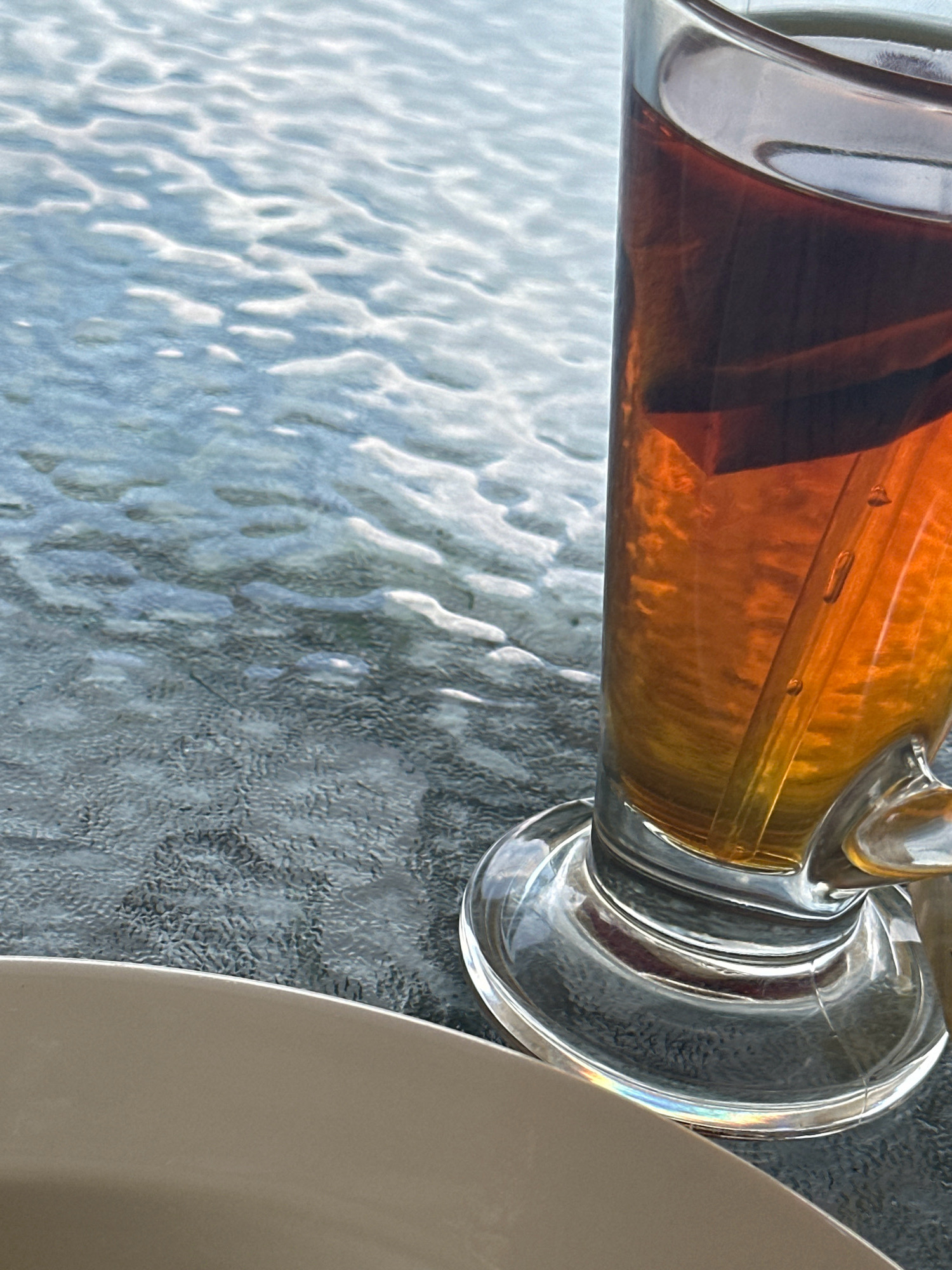 A close-up shot shows a glass of iced tea with a tea bag inside, placed on a textured surface that appears to be a glass table. The tea is a warm amber color and the glass has condensation. The background is blurred and shows a rippled pattern of light and shadow, suggesting an outdoor setting with water or a similar reflective surface. A portion of a light-colored plate is visible in the lower left corner of the frame. The lighting suggests daytime. The scene is devoid of people. The location is Kageshwori Manohara, Nepal.