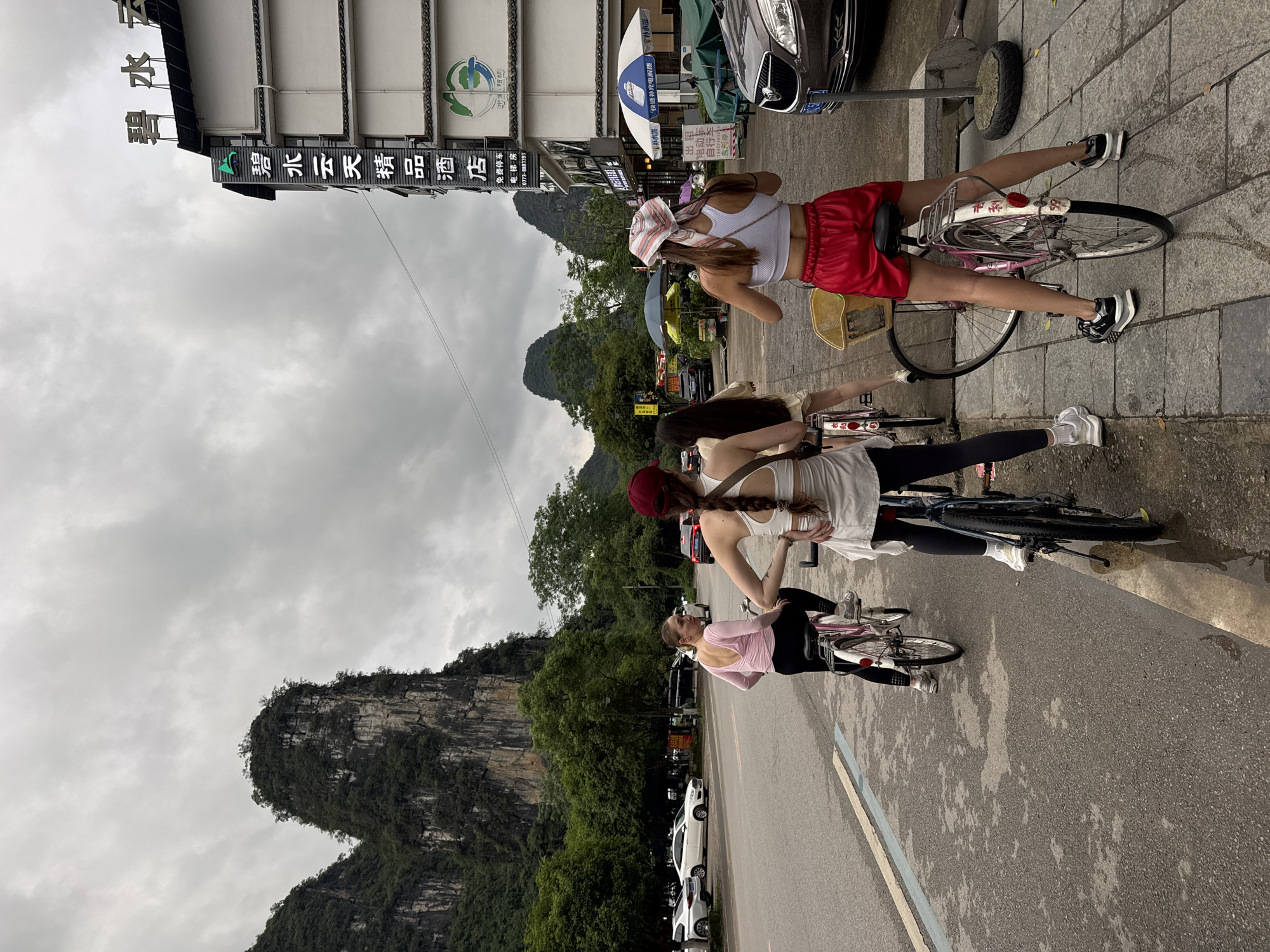 Three women are positioned on the side of a road with their bicycles in Yangshuo County, Guilin, China. In the background, a karst mountain is visible. The women are on a paved road next to a gray stone sidewalk. A car is parked next to a bus stop. The text on the building's signage includes the Chinese characters 碧水云 and the English words "electric car" and "free parking."