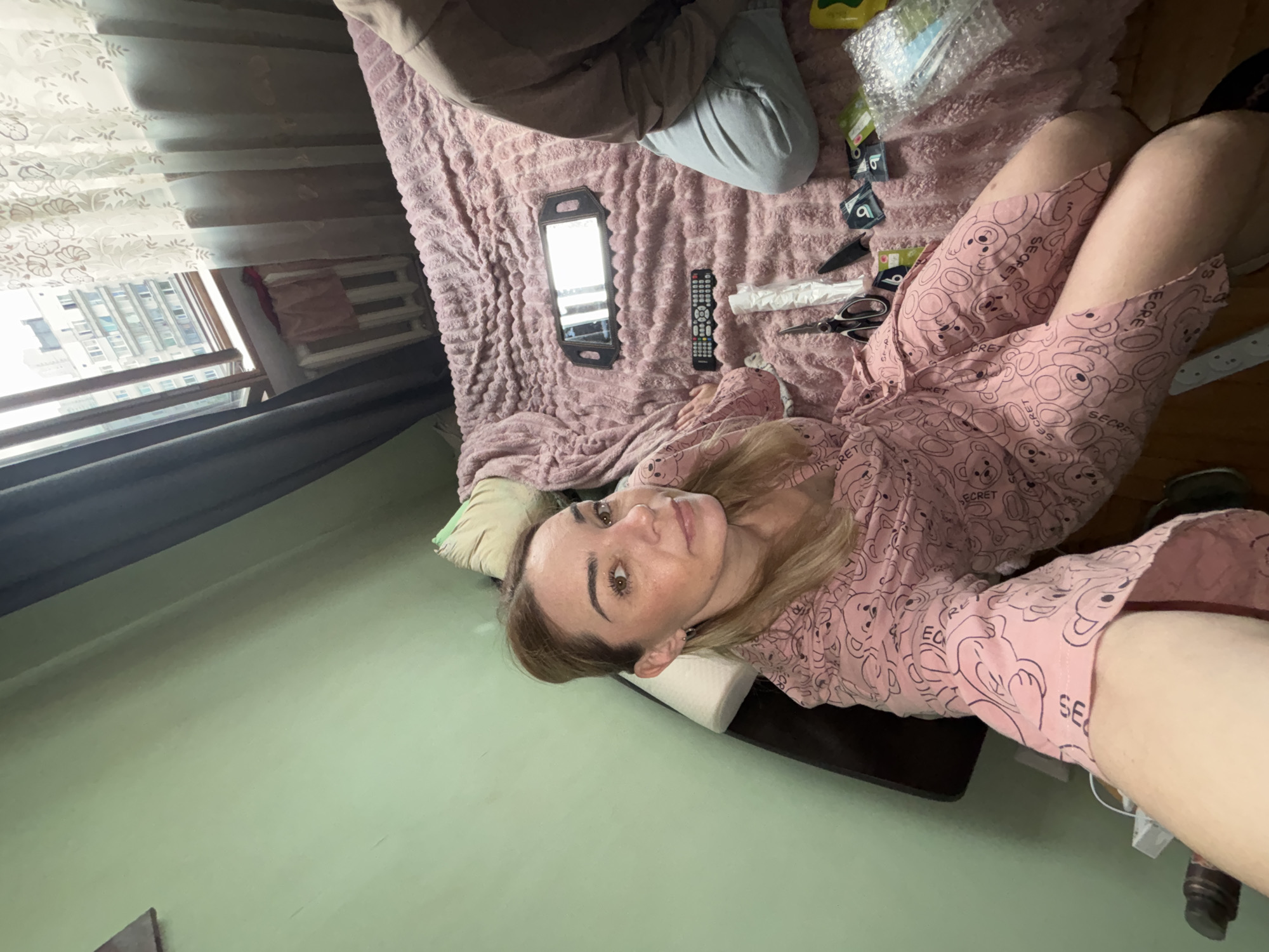 A woman with blonde hair pulled back, wearing a pink robe adorned with a pattern of cartoon bears and the word "SECRET" repeated, is lying on a bed. Her eyes are open, and she is looking at the camera. The bed is covered with a textured pink blanket. To her right, on the bed, are several objects including a small mirror with a dark frame, a television remote control, a pair of scissors, a knife, and several small packages that appear to be condoms. The room has light green walls. Through a window in the background, a multi-story building and a radiator are visible. A second person, wearing blue jeans and a grey top, is partially visible on the right side of the bed, also seated. The scene suggests a casual, indoor setting.