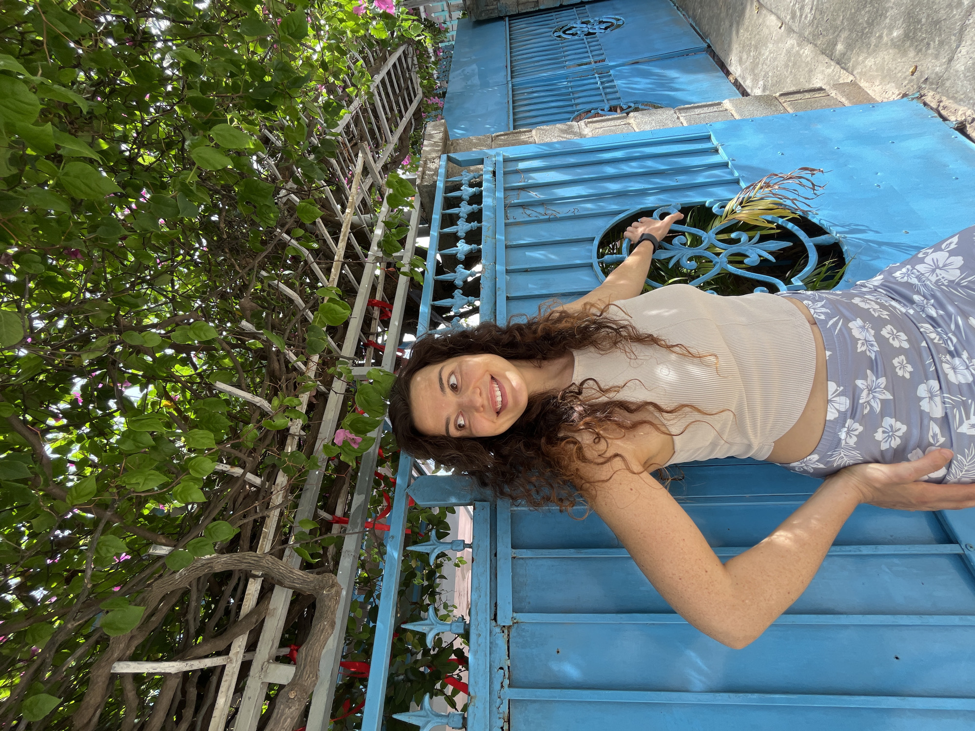 A woman with curly brown hair is standing in front of a bright blue gate in Mũi Né, Vietnam. She is wearing a beige ribbed tank top and a light blue skirt with a floral pattern. She is smiling and looking to her left, with her right hand resting on the gate. The gate is ornate, with decorative scrollwork and pointed finials. Behind the gate, there are green leaves and branches of a bougainvillea tree, with hints of pink flowers. The ground is paved with concrete. The scene appears to be outdoors on a sunny day.