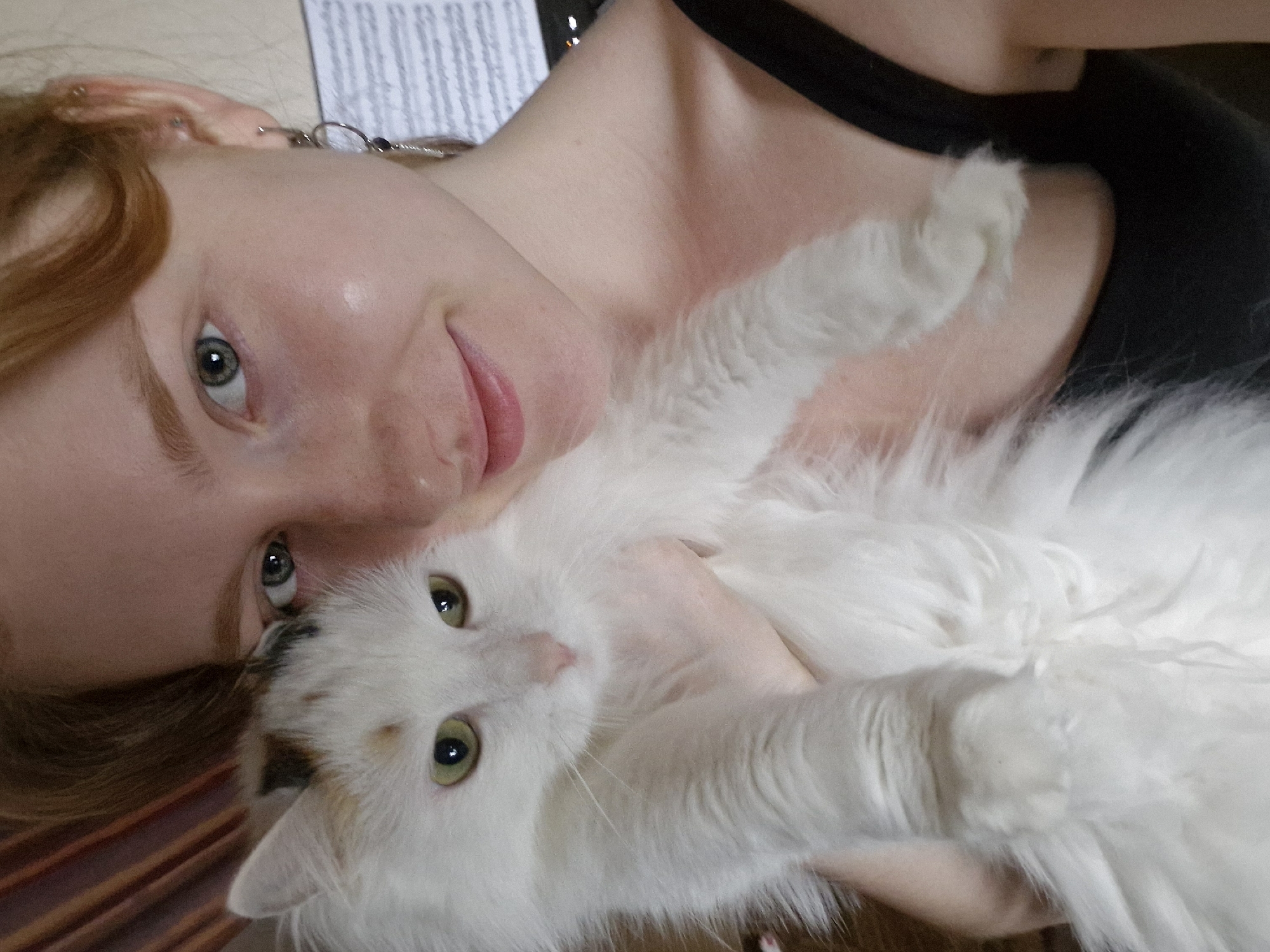 A person with red hair is lying down, holding a white, long-haired cat. The person is wearing a black tank top and earrings. The cat has yellow-green eyes. Part of a document is visible in the background. The location is not discernible from the image.