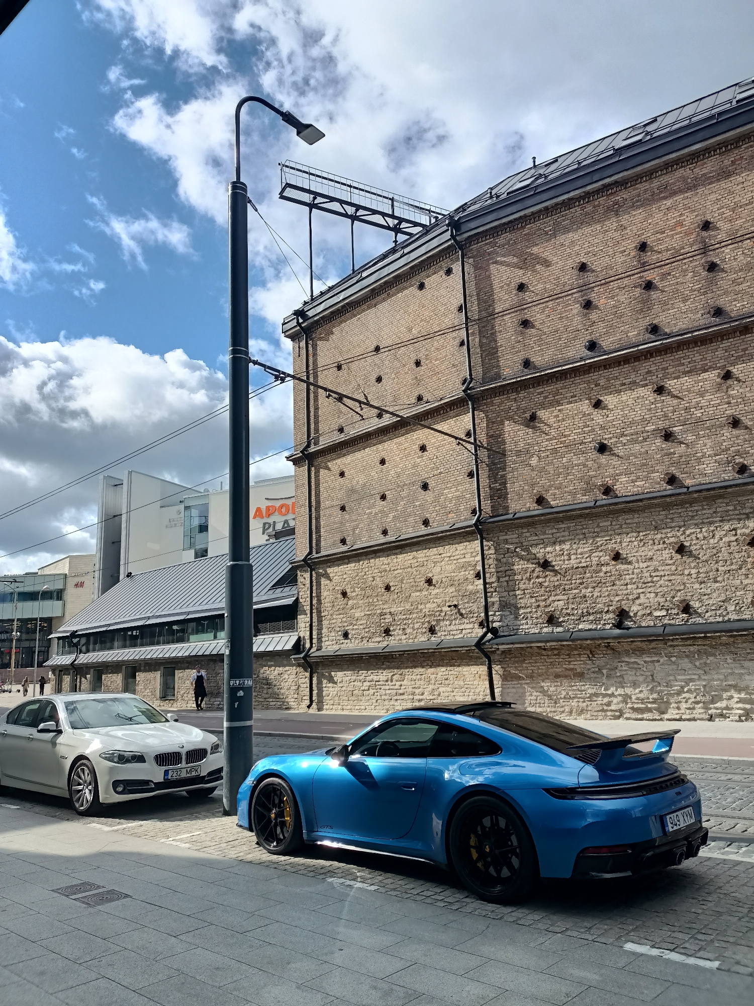 In Tallinn, Estonia, a bright blue Porsche 911 GTS is parked on a cobblestone street next to a white BMW sedan. The Porsche features a black spoiler and black wheels with yellow brake calipers. The BMW has a license plate that reads "232 MPK". Behind the cars, a tall brick building with protruding metal fixtures stands prominently. The street is lined with tram tracks. In the background, modern buildings, including one with an "APOLLO PLAZA" sign and an "H&M" store, are visible. The sky is partly cloudy with a blue sky and white clouds. A streetlamp stands to the left of the frame, obscuring part of the scene.