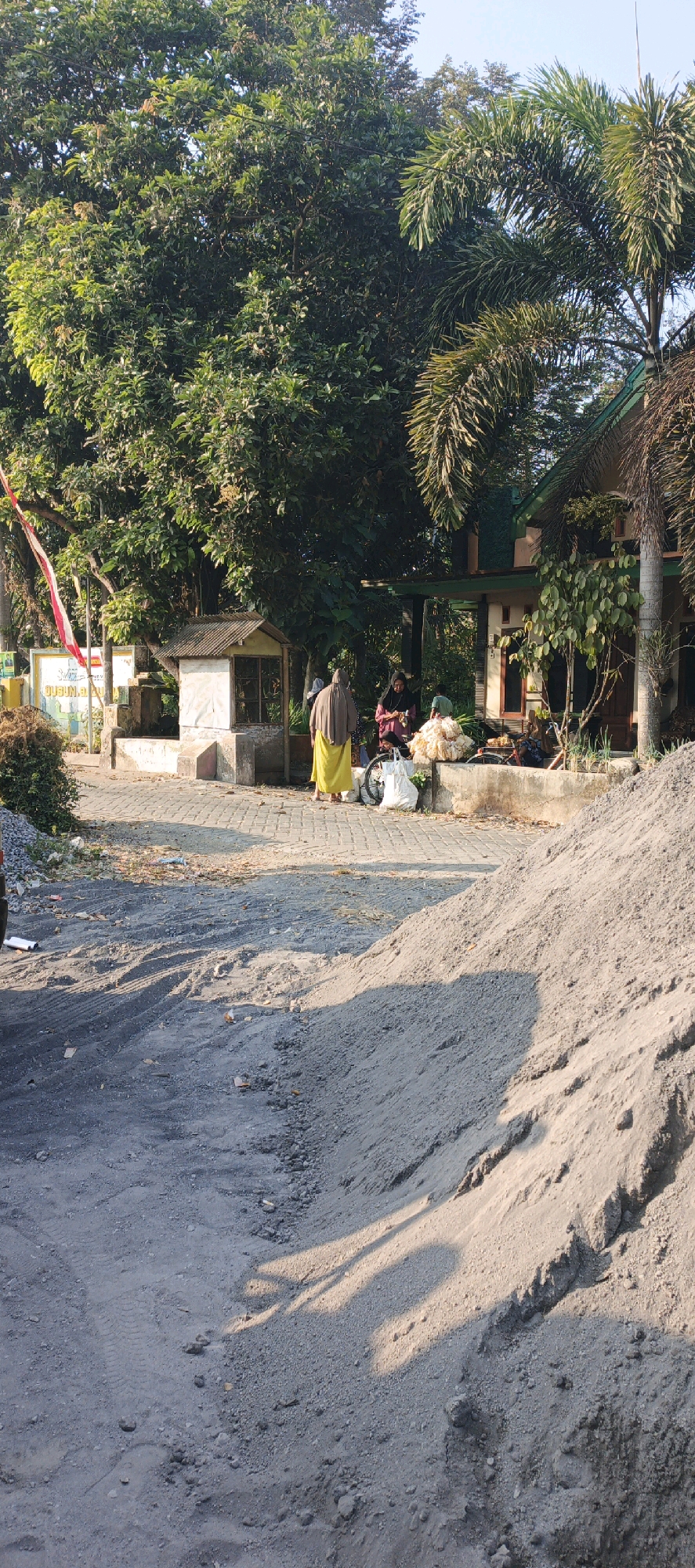 In Kasiyan, Indonesia, a roadside scene unfolds under a bright sky. A pile of grey sand sits to the right of a paved road. In the background, a building with a green roof is partially visible, surrounded by lush trees and palm fronds. Near the building, two women are interacting with a third person, possibly a child, who is out of frame. One woman is wearing a yellow robe and a headscarf, while the other is in a dark headscarf and a patterned dress. They are near a cart with a bicycle wheel. A small, shaded structure with a tin roof stands to the left, next to a yellow sign with red and white markings. A red and white flag flutters from a pole near the sign. The ground is a mixture of pavement and dirt, with some debris scattered on the left side of the road.