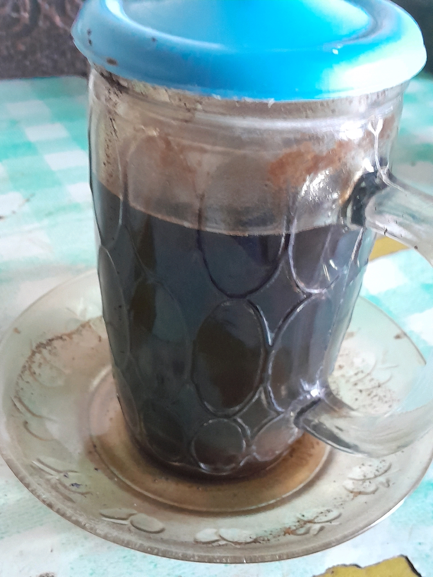 A glass mug filled with dark liquid, likely coffee, sits on a saucer. The mug has a handle and a blue plastic lid. The glass has a textured pattern of raised ovals. The saucer is clear glass with a decorative etched pattern. The mug and saucer are on a light-colored surface with a green and white checkered pattern. The background is blurred, but appears to show a textured surface. The scene suggests a casual setting, possibly a home or a small cafe in Glanggang, Indonesia.