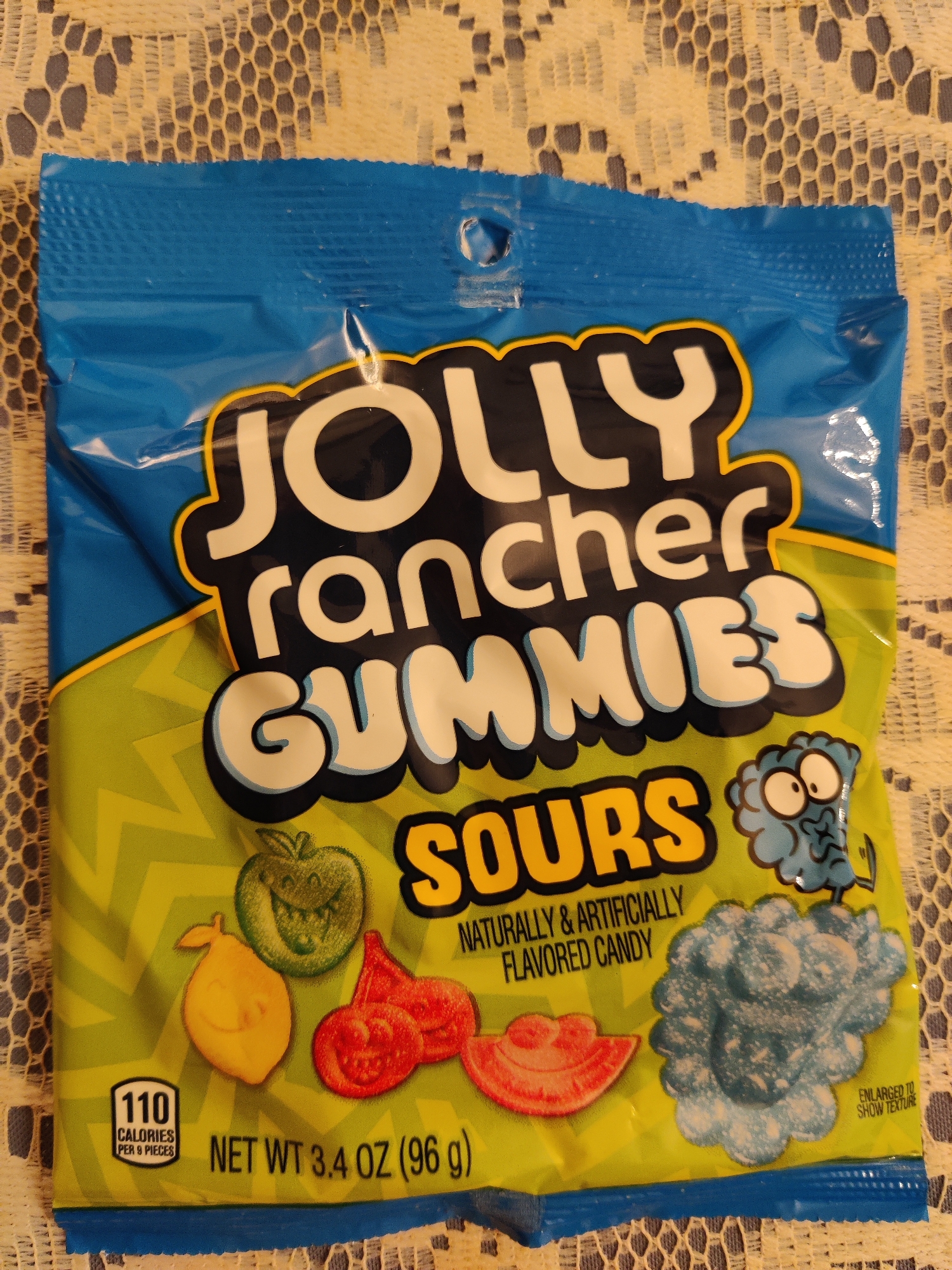 An overhead, medium shot captures a bag of Jolly Rancher Gummies Sours candies. The bag is predominantly blue at the top, transitioning to a lime green with a yellow zig-zag pattern towards the bottom. The Jolly Rancher logo is prominent, with "JOLLY" in large, white, stylized letters outlined in yellow and black, followed by "rancher" in smaller white letters. Below this, "GUMMIES" is written in large, white, puffy letters with a blue outline. The word "Sours" is in large, yellow, stylized letters outlined in black.
To the left of "Sours," an illustration of a green apple with a mischievous face is depicted, alongside a yellow pear-shaped candy. To the right, three red, circular candies with smiley faces are shown, and next to them is a cluster of blue, powdery candies, labeled "ENLARGED TO SHOW TEXTURE."
In the bottom left corner, a nutritional information icon displays "110 CALORIES PER 6 PIECES." Below this, the text "NET WT 3.4 OZ (96 g)" is visible. The bag rests on a white, lace-like tablecloth. The scene is located in Lower Paxton Township, United States of America.