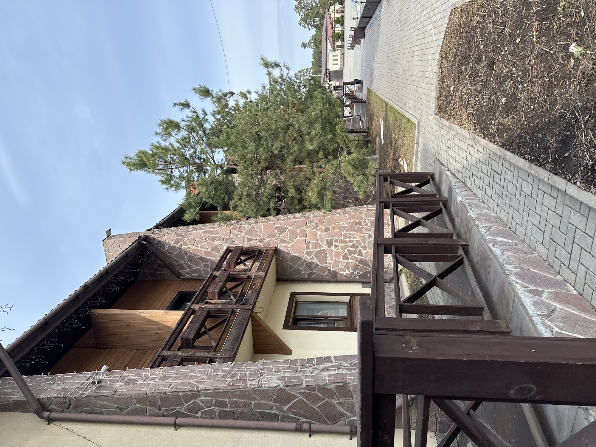 The image depicts a building with a stone and wood facade, located in an unidentified part of Votkinsk, Russia. The building appears to be a multi-story structure with a prominent stone gable. The roofline is decorated with string lights. A large pine tree partially obscures the upper portion of the building. A brown wooden railing extends along a stone-paved walkway adjacent to the building. The walkway leads toward other buildings in the background, suggesting a residential or commercial area. Adjacent to the walkway is a plot of bare earth. The sky is overcast with light blue hues. No people are visible in the frame.