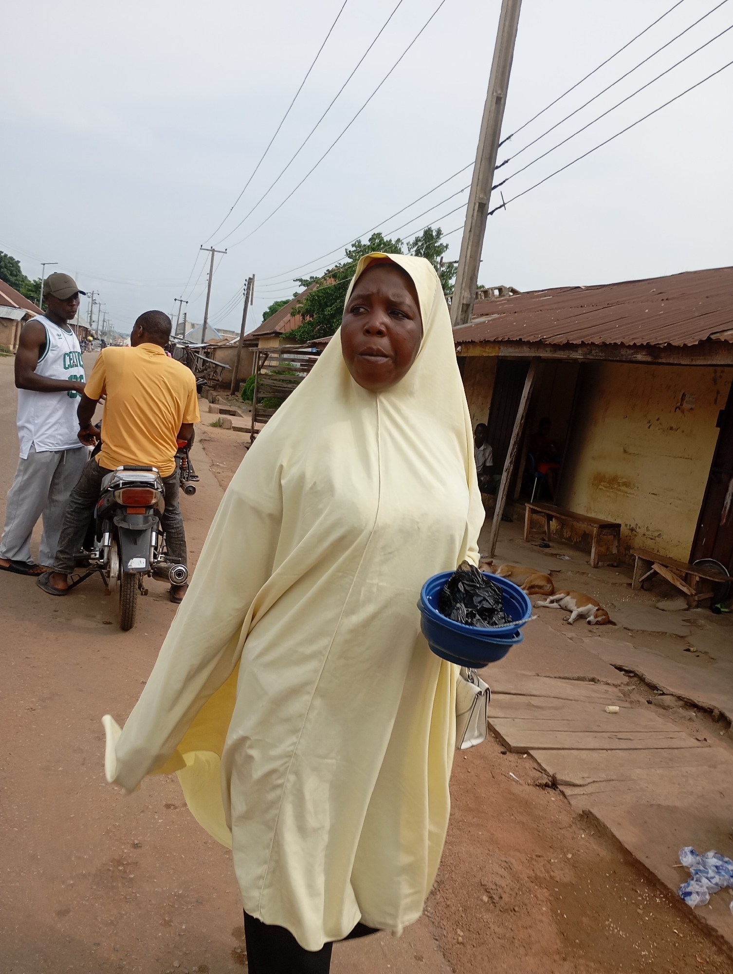 A woman wearing a light yellow hijab and a matching garment holds a blue bowl filled with black items, possibly food or refuse, in her right hand. She is walking on a dirt road in Shendam, Nigeria, alongside a building with a corrugated metal roof. Behind her, two men are on a motorcycle. One man, wearing a yellow t-shirt, is the driver, and the other, in a white tank top and a cap, is standing. Two dogs are lying down in the background. The scene appears to be a street or market area with basic buildings and overhead power lines. The overall atmosphere is one of everyday life in a Nigerian town.