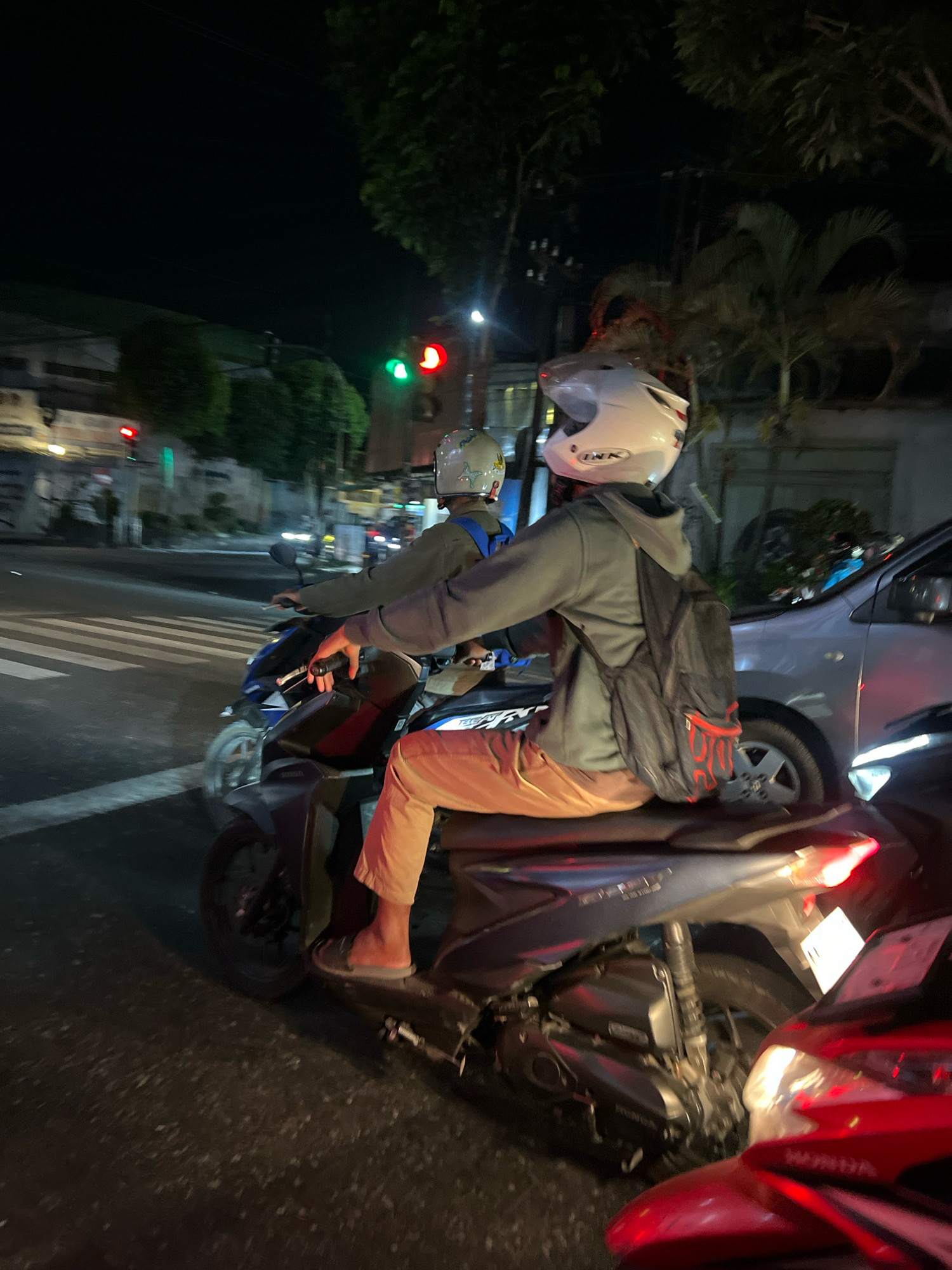 At night in Puncungrejo, Indonesia, two individuals are riding a motorcycle. The rider in front wears a white helmet and a light-colored jacket. The passenger behind him, wearing a white helmet, has a backpack with red accents and is wearing tan pants and sandals. They are in a traffic intersection, with a traffic light visible showing a green light for one direction and a red light for another. The road surface is asphalt with pedestrian crossing markings. Other vehicles, including a silver car and a red motorcycle, are also present in the vicinity. The surrounding environment includes trees and buildings, suggesting an urban or suburban setting.