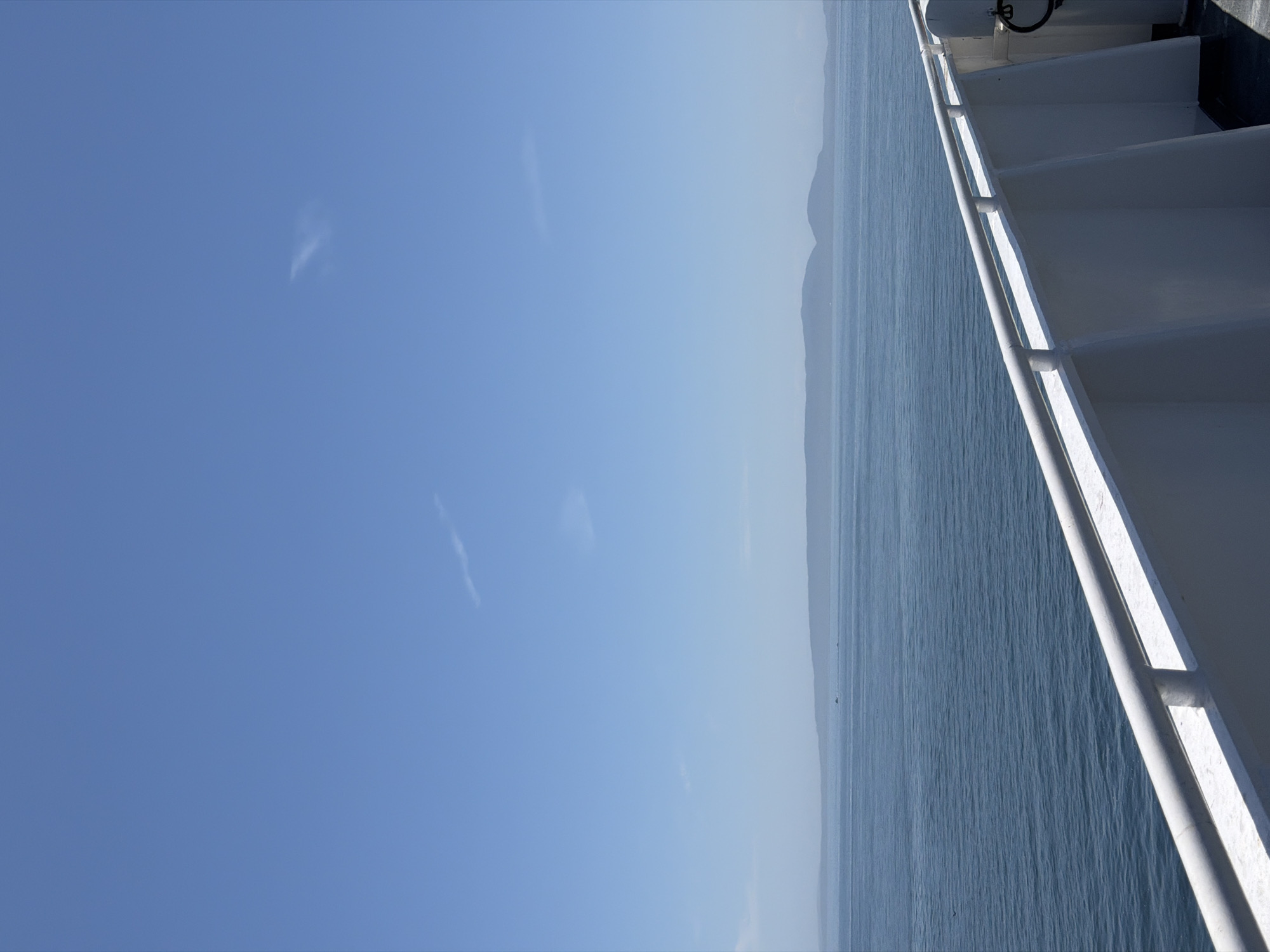 The image captures a serene view from a ferry deck, looking out over the Strait of Georgia. The vast expanse of calm, blue water dominates the lower two-thirds of the frame, with a distant, hazy shoreline of islands and mountains forming the horizon. A white railing runs along the right side of the image, indicating the vantage point is from the side of a vessel. The sky above is a clear, bright blue, with a few wispy clouds scattered sparsely. The overall impression is one of peaceful travel and natural beauty. This scene is likely captured while traveling on a BC Ferries vessel between Tsawwassen in Delta, Canada, and one of the Vancouver Island destinations.
