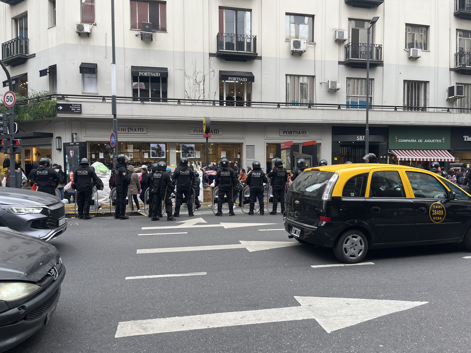 The image shows a street scene in Buenos Aires, Argentina, with a line of police officers in riot gear facing a crowd of people. Behind the officers, there are buildings with storefronts, including a store named "PORTSAID" and a toy store called "COMPAÑÍA DE JUGUETES". There are several cars in the foreground, including a black and yellow taxi with the license plate "KOF 902" and the taxi number "38489 GCBA." The road has a white arrow indicating the direction of traffic. The street sign indicates Avenida Corrientes. The image suggests a demonstration or gathering taking place.
