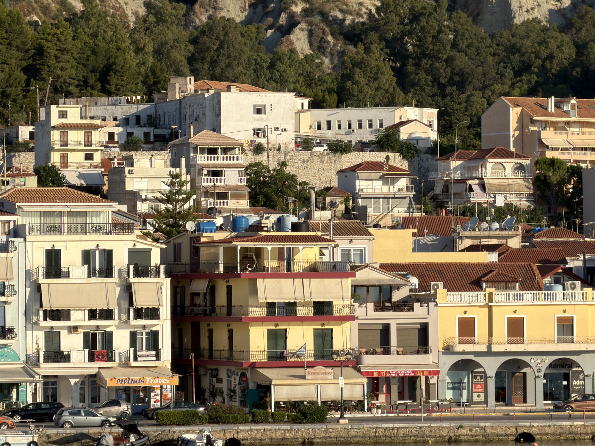 The image depicts a waterfront scene in Zakynthos Municipal Unit, Greece, characterized by a dense collection of multi-story buildings lining a harbor. The architecture is a mix of traditional and more modern styles, with many buildings featuring balconies, awnings, and red-tiled roofs. Some buildings are painted in bright colors like yellow and cream, while others are a more muted white or grey.
In the foreground, a waterfront promenade runs along the edge of the harbor. Boats are visible docked in the water, and several small inflatables are tied to a dock. The promenade is lined with a variety of businesses, including shops and restaurants, indicated by signage such as "Mythos Travel" and "Famous Bamboo Chinese Restaurant". Cars are parked along the street in front of these establishments.
Behind the waterfront buildings, the terrain rises sharply, covered in dense green trees and a rocky, light-colored cliff face. The sunlight suggests it is either morning or late afternoon, casting distinct shadows and highlighting the textures of the buildings and the natural landscape.
There are no prominent individuals in the scene. The focus is on the urban and natural environment, showcasing the architectural style and the setting of this coastal town. The overall impression is one of a bustling, sunlit coastal settlement.