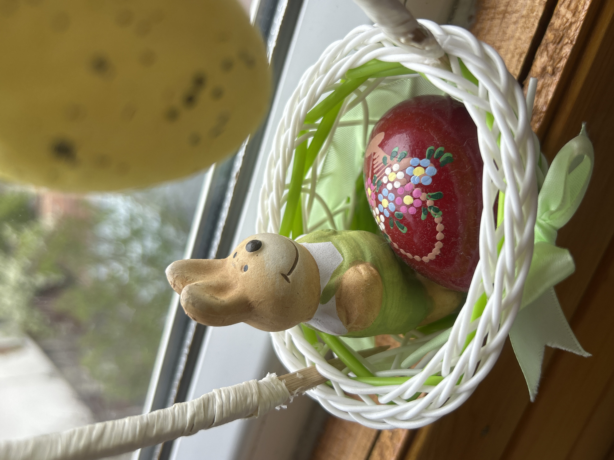 Here is a close-up shot of an Easter-themed decorative arrangement. A woven white basket with green accents and a light green ribbon sits near a window. Inside the basket is a red Easter egg decorated with a floral design. Next to the egg is a small, ceramic duck figure. A yellow, spotted object, possibly another Easter egg, hangs partially visible in the upper left corner of the frame. The setting appears to be indoors, near a window, judging by the outdoor scene visible through the glass.