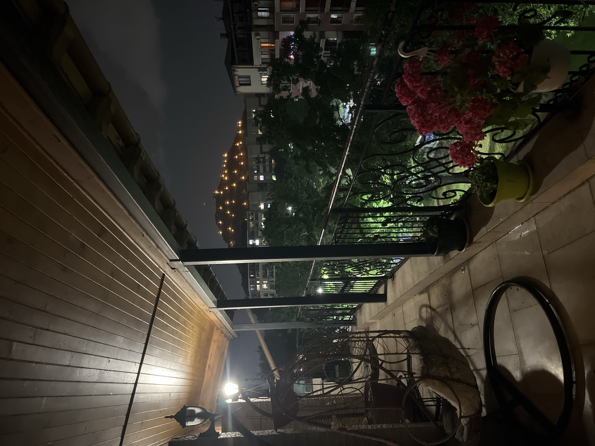 This image presents a nighttime view from a covered balcony overlooking an urban landscape identified as Plovdiv, Bulgaria. The balcony features a horizontal wooden plank ceiling, illuminated by an antique-style dark metal wall lantern positioned on the left. Below the lantern, a secondary light source casts a bright glow on the immediate surroundings. A dark-colored, possibly wicker, hanging swing chair with a light-colored cushion is situated on the left side of the balcony. The balcony floor is composed of light-colored, square tiles that exhibit some reflective areas. A black wrought-iron railing with ornate scrollwork extends along the perimeter of the balcony, supported by dark vertical posts.
On the right side of the balcony, a white hanging pot containing numerous red flowering plants, likely geraniums, is affixed to the railing. Adjacent to it, a smaller yellow pot with a green plant rests on the tiled floor.
Beyond the balcony, a canopy of dark trees is visible, interspersed with multi-story residential buildings that display illuminated windows, indicating occupancy. In the background, a prominent hill is clearly discernible, covered with numerous small, distinct light sources evenly distributed across its slopes. This illuminated geographical feature is characteristic of one of the historical hills of Plovdiv, Bulgaria, such as Nebet Tepe. The night sky above is dark, with indistinct cloud formations visible. No human figures are present in the scene.