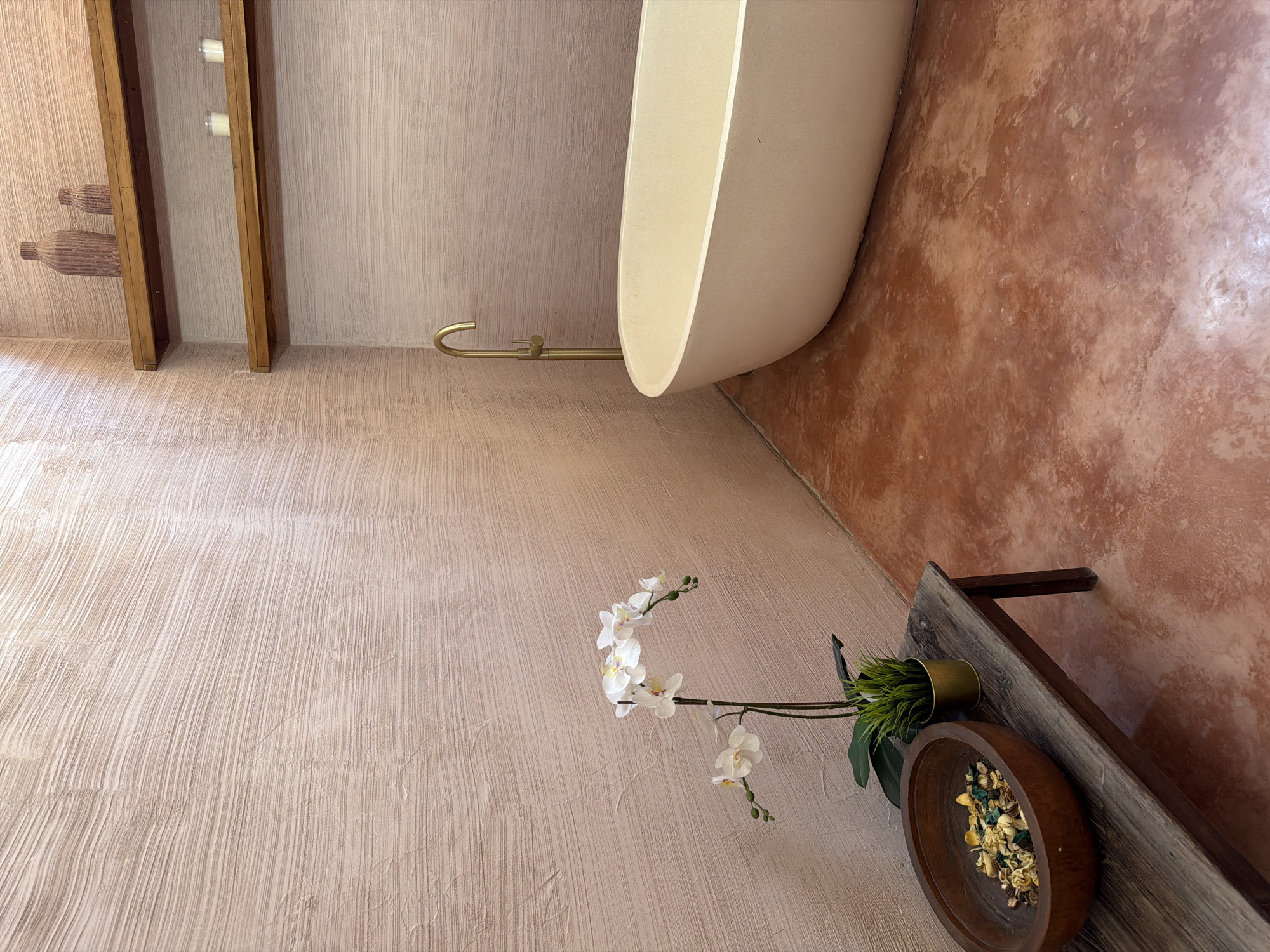An upward-angled, interior shot reveals a bathroom. The space features textured walls, light beige on one side and terracotta on the other. A freestanding, light-colored bathtub is positioned between the two differently colored walls, with a gold-colored faucet. A wooden shelf with candles and decorative objects is attached to the lighter beige wall, while another wooden shelf, holding a pot of greenery and a wooden bowl filled with potpourri, is attached to the terracotta wall. A sprig of white orchids is placed near the pot of greenery. The location is North Kuta, Indonesia.