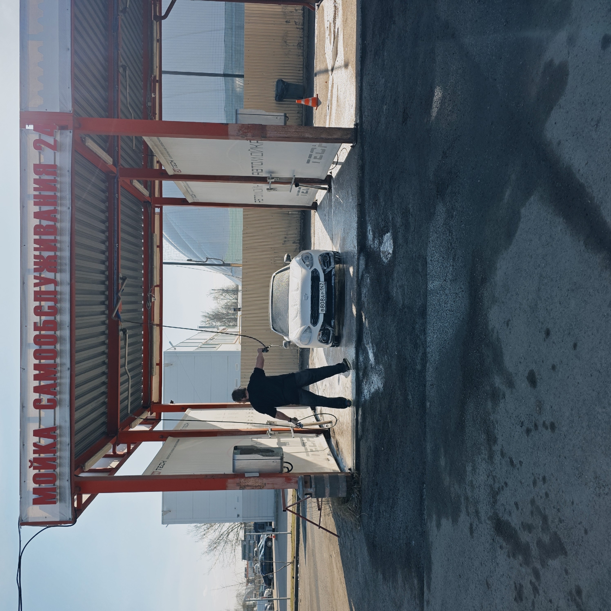 The image depicts a self-service car wash in Saint Petersburg, Russia. A man dressed in black is actively washing a white Fiat 500 car. The car's license plate is partially visible, reading "B 808 AP 177." The car wash structure features a red metal frame with a sign in Russian, "МОЙКА САМООБСЛУЖИВАНИЯ 24." The ground is wet, indicating recent or ongoing washing activity. In the background, there's a building with reflective windows, a traffic cone, and other parked vehicles visible. The sky is clear and bright, suggesting a sunny day.