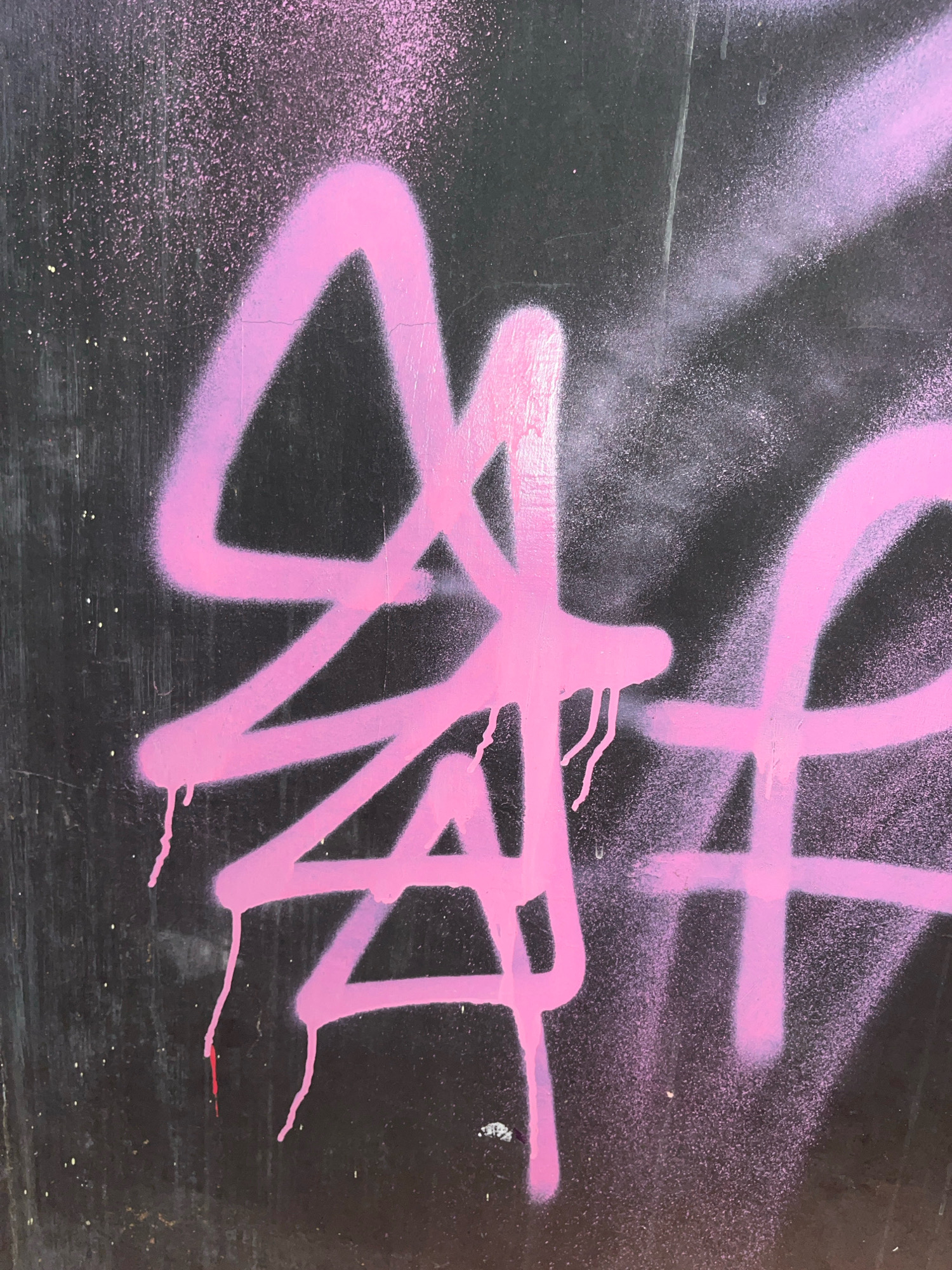 A close-up, full-frame shot shows pink spray paint graffiti on a dark, textured surface. The graffiti consists of thick, abstract lines and splatters, with drips visible on some of the strokes. The background appears to be a weathered, possibly metallic surface, with some light speckles. The lighting is even, highlighting the texture of the paint and the surface. The location in the city of Surakarta, Indonesia, is not visually identifiable from the image. There are no people, actions, or interactions depicted.