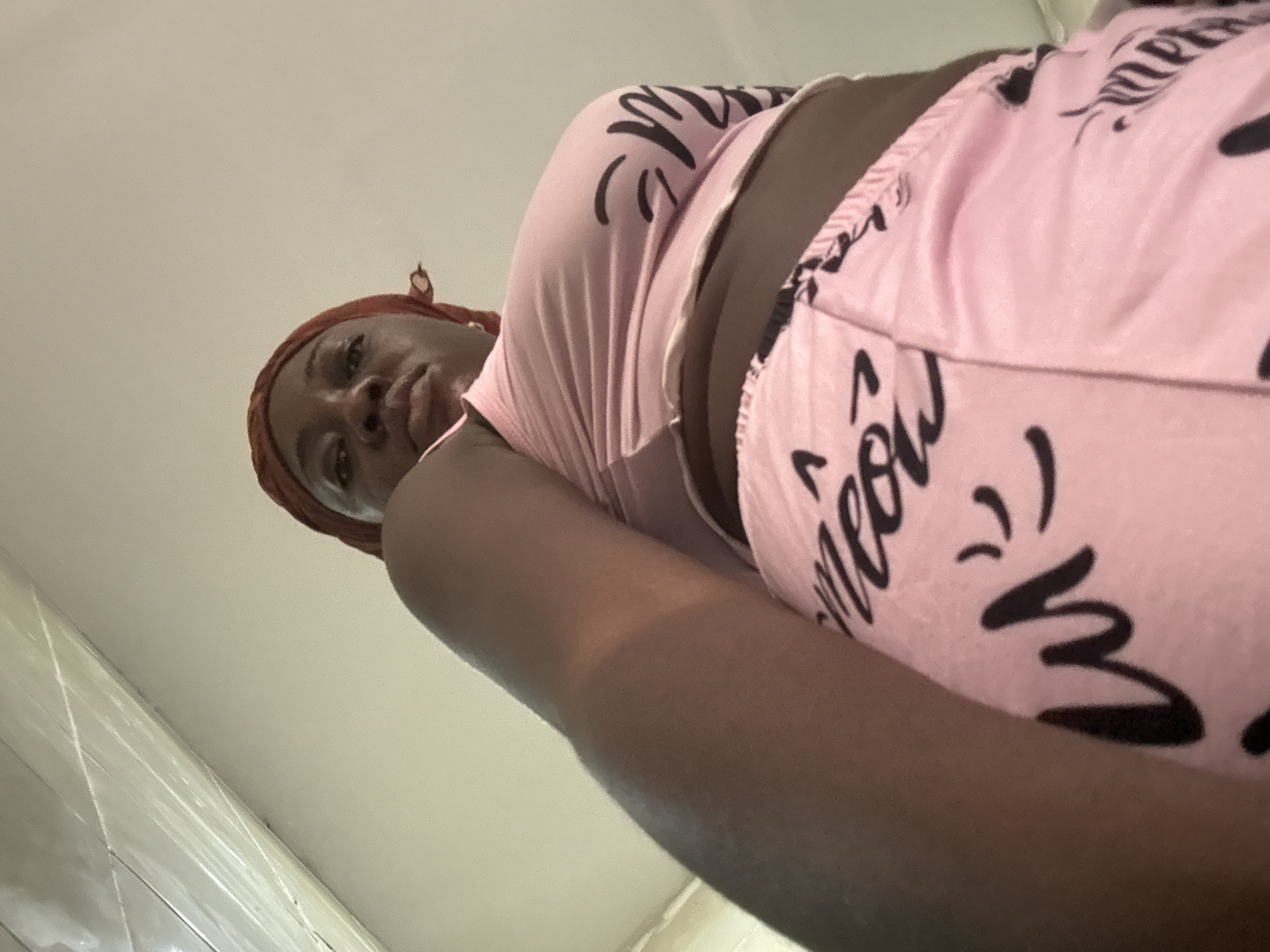 A low-angle, upward-facing shot captures a person looking down towards the camera in an indoor setting. The individual, who has dark skin, is positioned in the upper right portion of the frame. They wear a reddish-brown head covering and a light pink two-piece outfit adorned with black stylized text and graphics.
The pink attire features the word "meow" repeated in a playful, handwritten font, along with what appear to be cat whiskers or paw print designs. The person's face is partially visible, showing dark eyes and lips, with a neutral expression as they gaze directly downwards. Their body is angled, and their left arm extends into the lower right of the frame.
The background primarily consists of a plain, light-colored wall or ceiling. In the lower left corner, a section of white or light-colored glossy tiles with horizontal lines is visible, suggesting a tiled floor or lower wall area, possibly within a bathroom or kitchen. The extreme low perspective implies the camera is placed on the ground or a very low surface, with the person bending or looking over it. The time of day is not discernible, and the setting is clearly indoors. The overall context suggests a casual, perhaps self-taken, photograph within a home in Iddo Tudunwada, Nigeria.