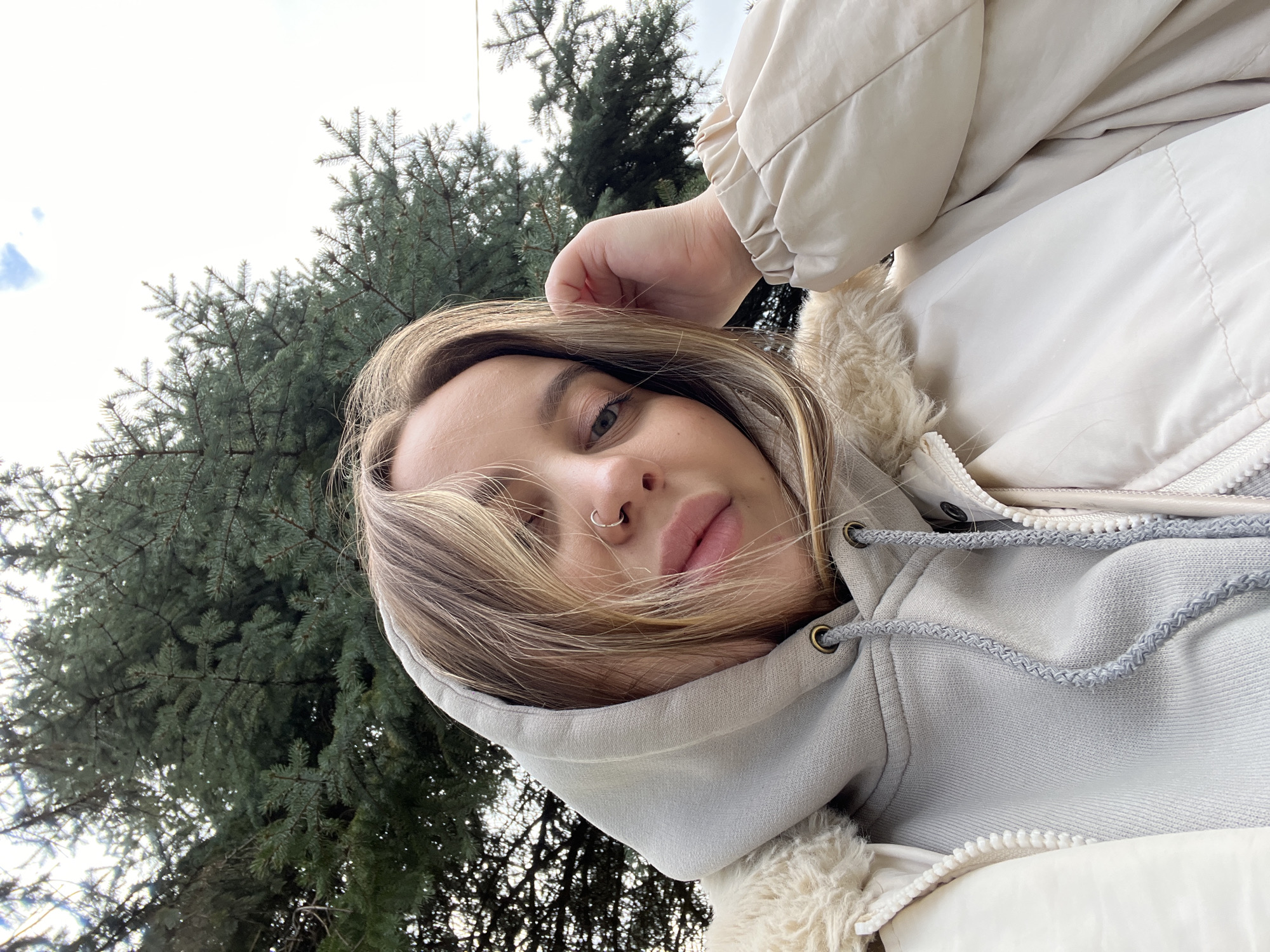 A young woman poses outdoors, under what appears to be a pine tree, looking down at the camera. She has blonde hair, a nose ring, and wears a hooded sweatshirt underneath a cream-colored jacket. The location is undetermined; there is no evidence to connect the image to the city of Novopokrovskaya, Russia.