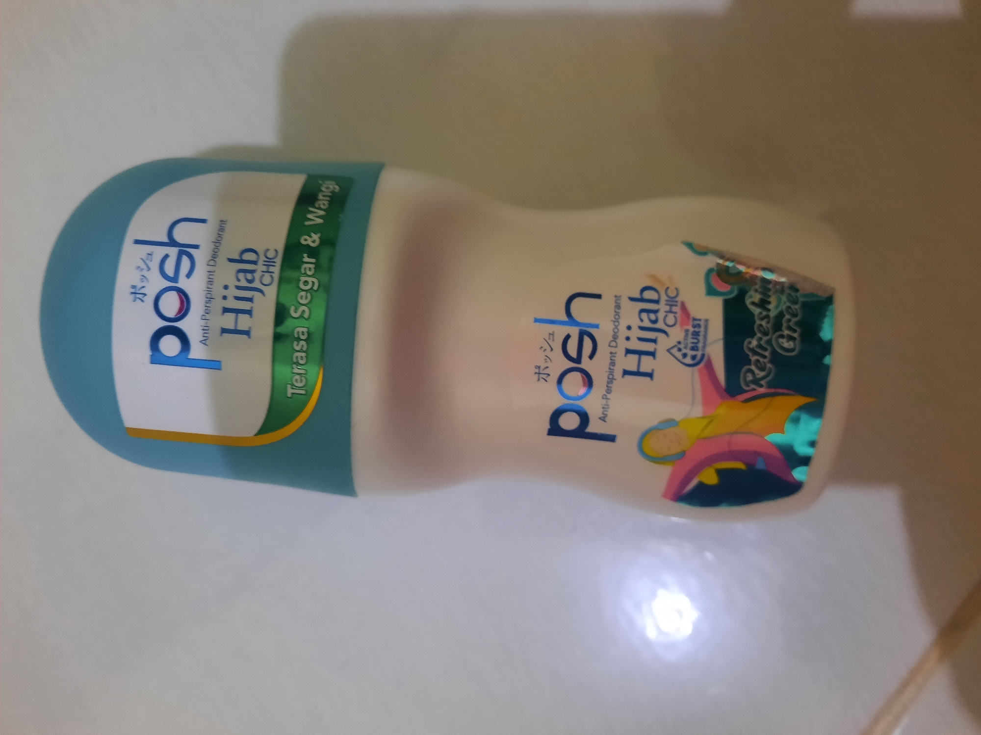 A Posh Hijab Chic anti-perspirant deodorant is photographed indoors. The deodorant is a white bottle with a teal blue cap. The label on the bottle has Japanese characters, "posh," "Anti-Perspirant Deodorant," "Hijab CHIC," and "Terasa Segar & Wangi." There is also a graphic of a person wearing a hijab and headphones on the lower portion of the label, with the words "ACTIVE BURST FRAGRANCE" and "Refreshing Green." The background is a blurred white wall. There is no indication of the city of Lhokseumawe, Indonesia.
