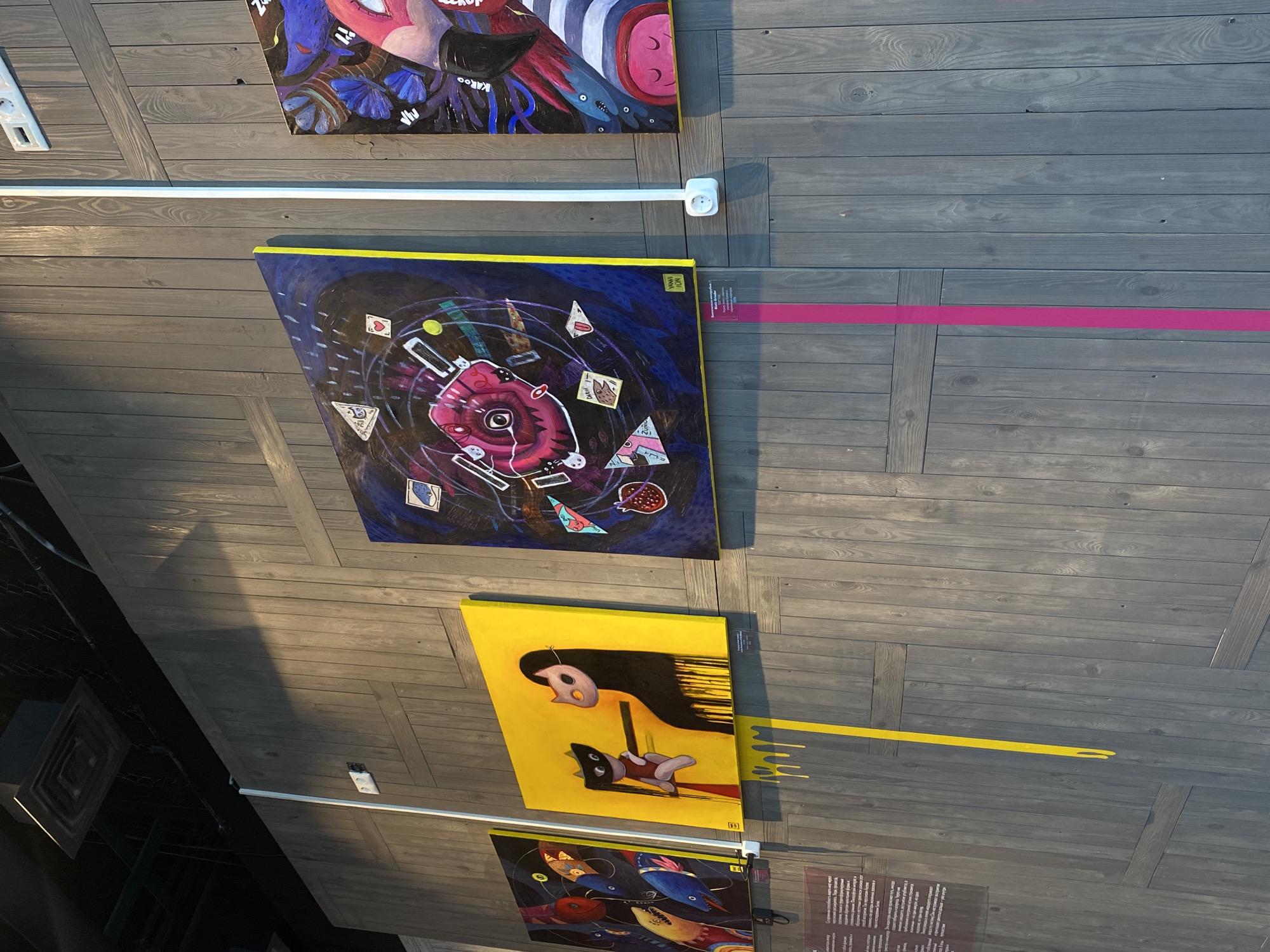 The image shows an indoor space, likely a gallery or museum, featuring several framed paintings hanging on a dark gray wooden wall. The perspective is slightly angled, looking upwards at the wall.
From top to bottom, the paintings vary in style: the top one is abstract with vibrant colors; the second appears to be a surrealistic piece with darker colors and abstract shapes; the third one is a yellow painting depicting a figure with a mask-like face; and the bottom painting features multiple colorful bird-like creatures.
A white light fixture runs horizontally between the top two paintings. A pink stripe is visible on the wall next to the second painting. A yellow stripe is on the wall next to the third painting. Underneath the paintings are what appear to be informational cards or descriptions for each artwork.
Given the reference to Perm, Russia, this is likely taken in a gallery or exhibition space in Perm, showcasing contemporary art.