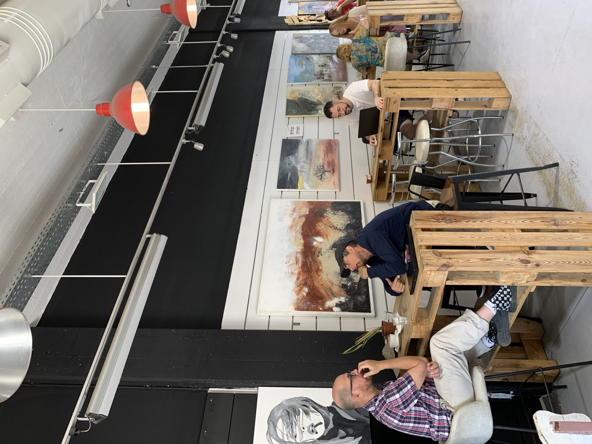The scene captures an interior space in what appears to be a collaborative working environment, possibly in Tel-Aviv, Israel, given the Hebrew text visible on one of the wall-mounted art pieces. The space is illuminated by a combination of natural light and industrial-style overhead lighting fixtures, some with red dome shades.
Several individuals are present, engaged in work or discussion. Desks constructed from wooden pallets provide workstations for these people. A man in a plaid shirt sits in a chair, appearing to be deep in thought or conversation, while a man in a navy shirt and baseball cap is leaning over a pallet desk and seems to be talking with the man in the plaid shirt. Another man in a white shirt is working at a computer. Other individuals can be seen in the background, at other desks.
The decor includes an eclectic collection of artwork hung along the walls, adding character to the space. The walls are painted in black and white stripes, which creates a dynamic visual element in the room. The flooring is a light-colored concrete or similar material.