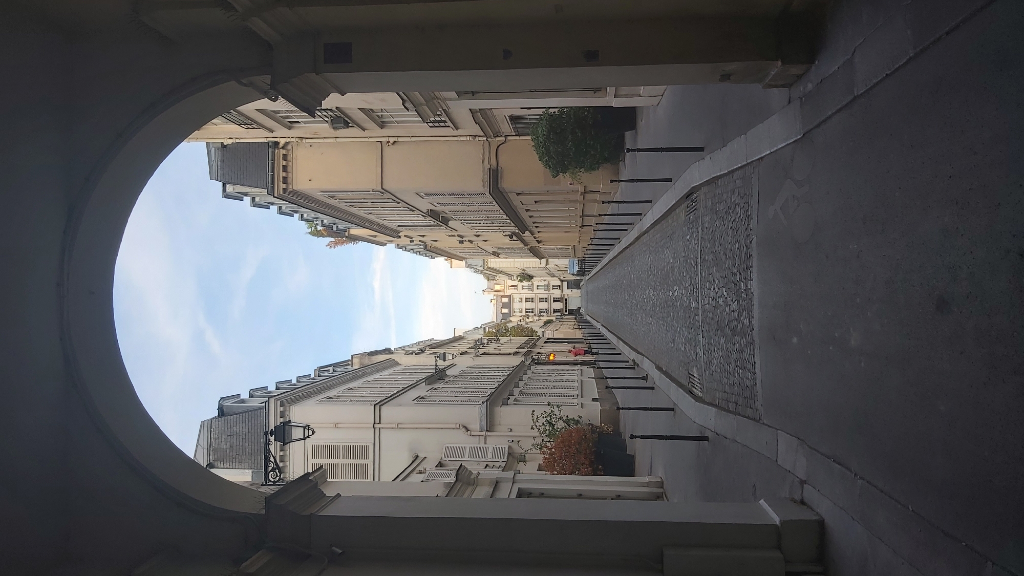 The image shows a view looking down a cobblestone street in Paris, France, framed by an archway. The street is lined with tall, cream-colored buildings with mansard roofs and numerous shuttered windows. A few trees are visible, some in planters on the sidewalk. The sky is light blue with some wispy clouds. At the end of the street, more buildings are visible. There is a single person walking down the street in the distance. The scene appears to be peaceful and quiet.