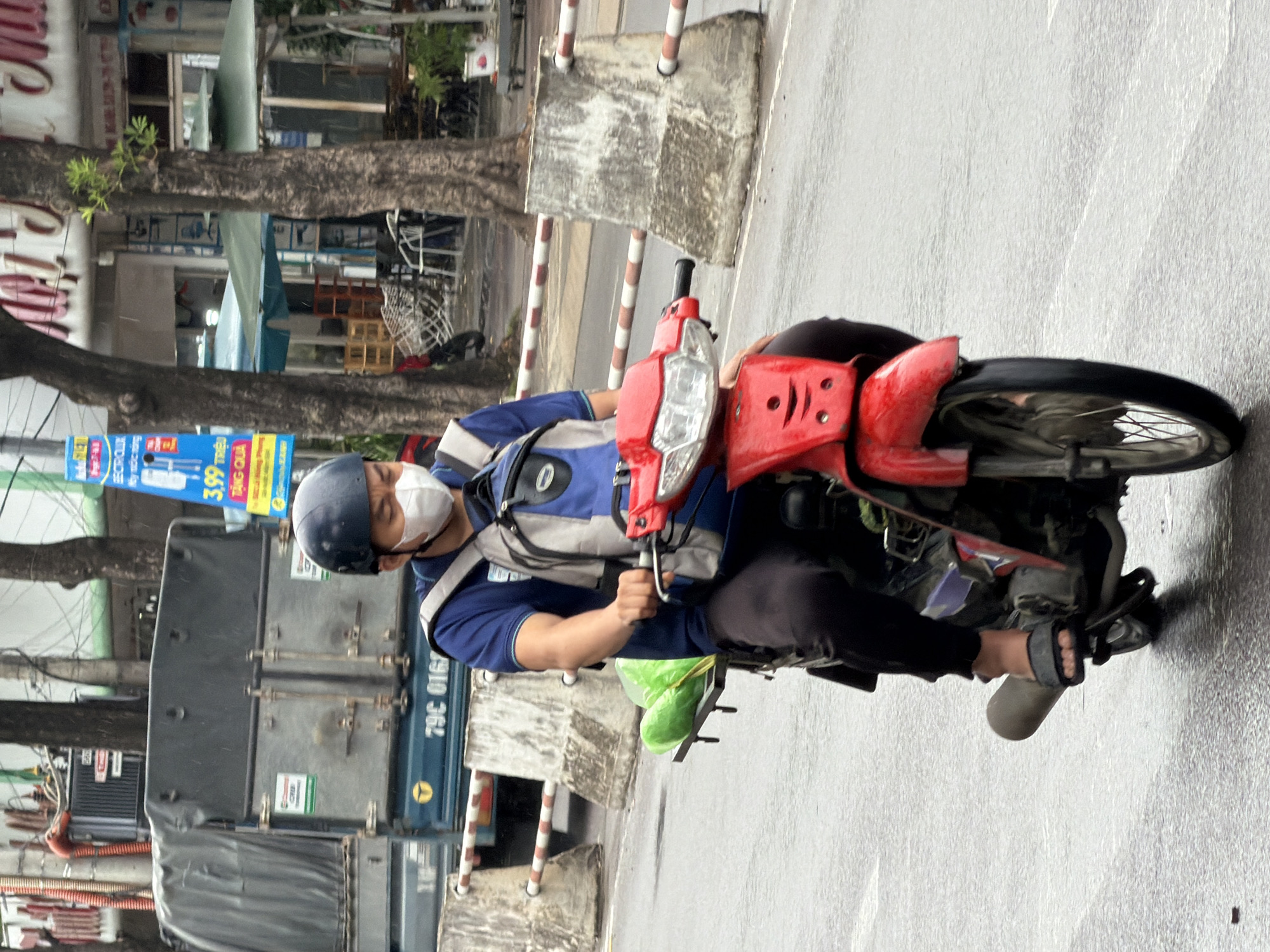 A person wearing a blue shirt, black pants, a dark blue helmet, and a white face mask rides a red motorcycle on a wet city street. The person has a backpack on, with a blue and grey design. Two large green plastic bags are attached to the back of the motorcycle. The background shows trees, buildings, and a parked truck. A blue and yellow sign with Vietnamese text is visible on a pole. The street appears to be in Nha Trang, Vietnam.
