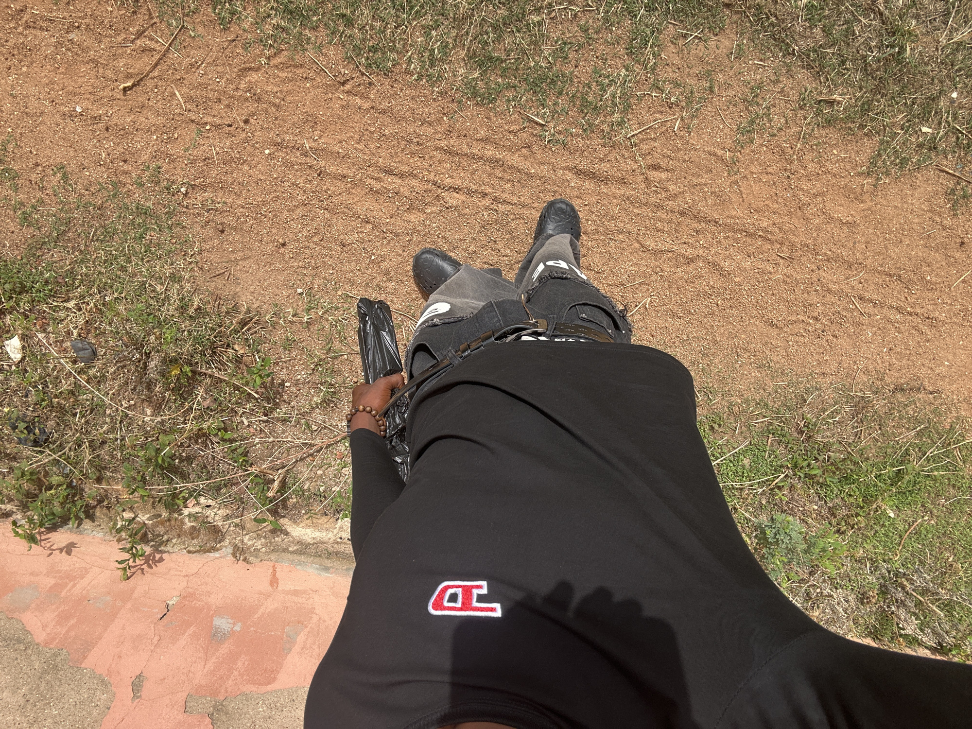 A person wearing black crocs, grey jeans, and a black long-sleeved shirt with a white and red logo is standing on a patch of dirt and grass in Kirana, Nigeria. The person is holding a black plastic bag in their left hand. The ground is a mixture of dirt, grass, and some concrete paving.