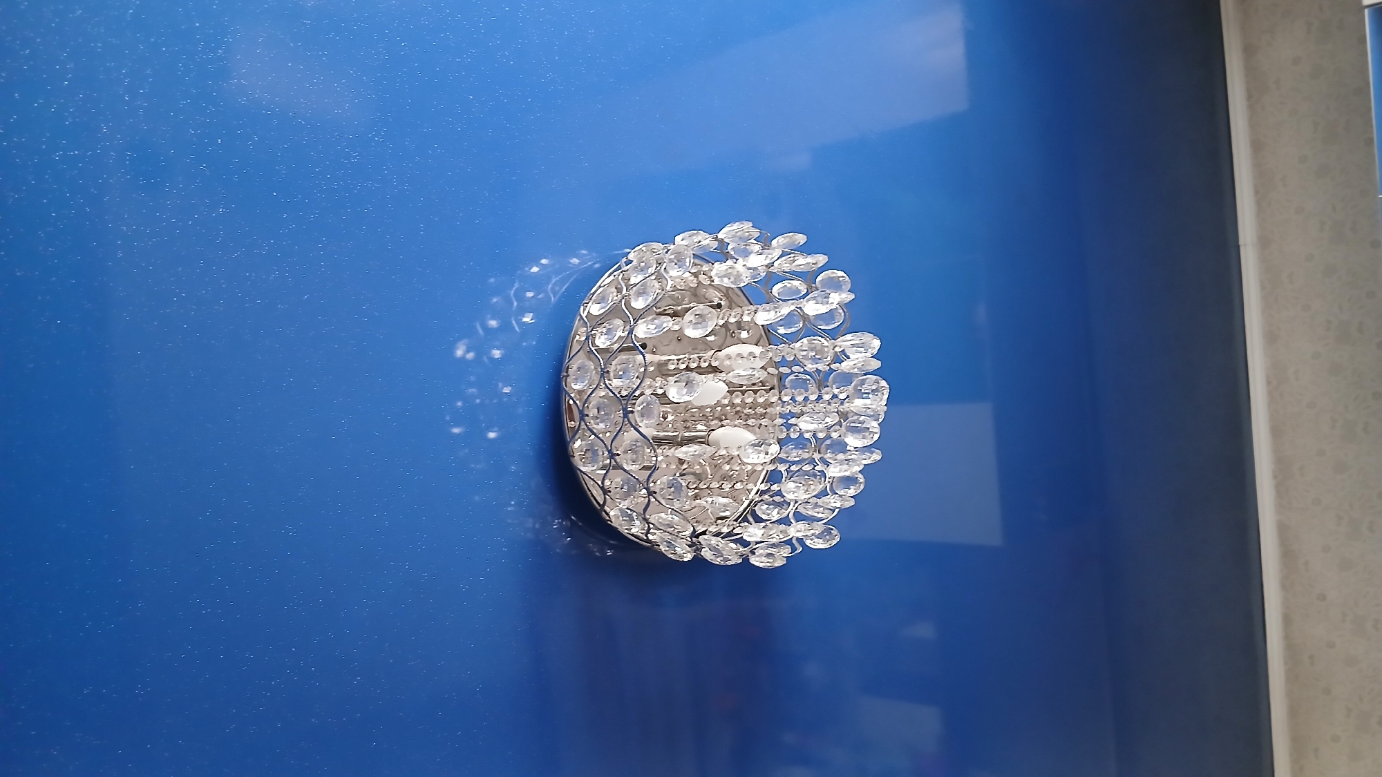 A crystal chandelier hangs from a blue, reflective ceiling in a room, likely a bedroom or living area. The chandelier is ornate, with many faceted crystal pendants that catch and refract light. The ceiling appears to be made of a glossy material, possibly stretch ceiling material, which reflects the light and the objects in the room. The lower portion of the image shows a textured wall or another surface, possibly a wallpapered wall. The scene is indoors and the lighting suggests it might be daytime, with the chandelier illuminated by ambient light. The location is identified as Stepnoye, Kyrgyzstan.