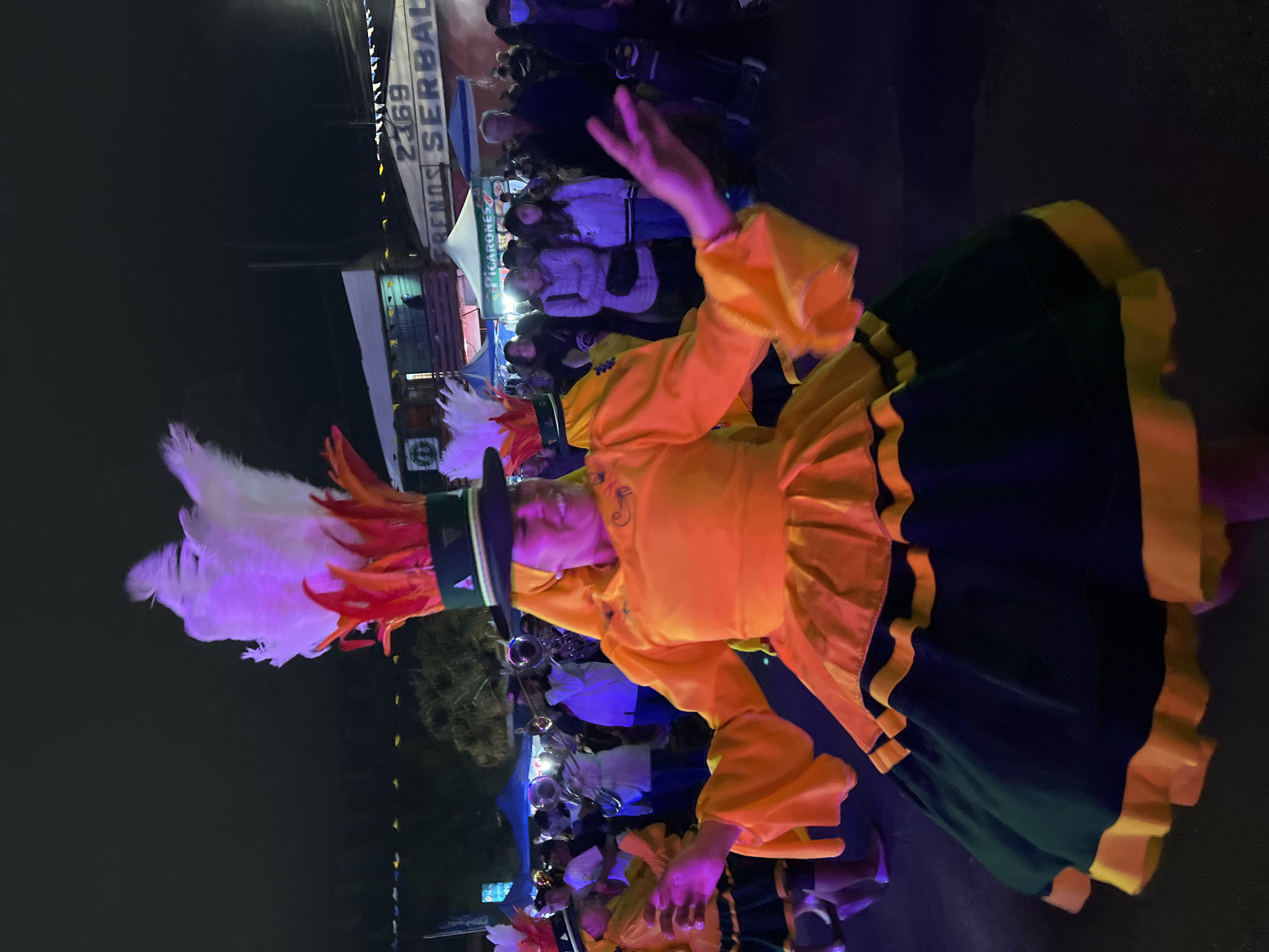 In the city of Arica, Chile, a woman in a vibrant, traditional orange and dark green costume with yellow trim dances at night. Her costume features voluminous sleeves and a layered skirt adorned with bands of yellow and black. She wears a tall, festive hat adorned with large white and red feathers, topped with a dark green band. Her face is lit from the side, and she smiles as she extends her right arm, palm open. Behind her, other performers and festival-goers are visible. A brass band is playing in the background, with some members holding instruments. Tents and stalls with signage are set up, suggesting a street fair or festival atmosphere. The lighting is dim, characteristic of an evening event, with artificial lights illuminating the scene. The background also shows some trees and darker, unlit areas. The ground appears to be asphalt or a similar paved surface. The overall impression is of a lively cultural celebration.
