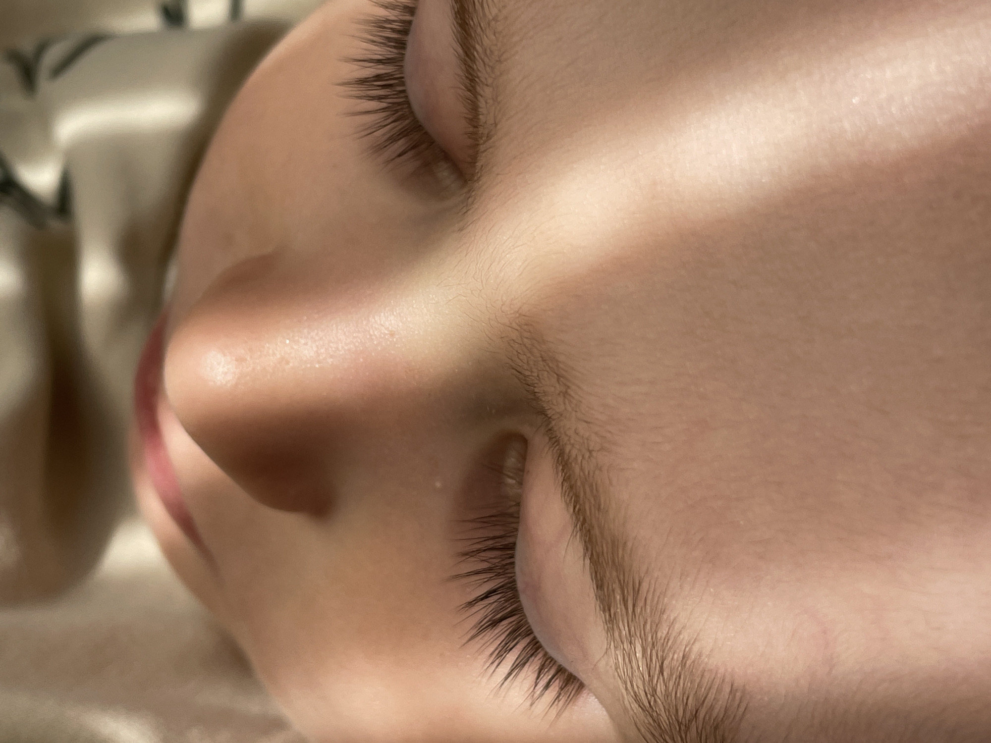 The image, described as located in the city of Lukintsy, Russia, presents a detailed, vertical close-up of a young individual's face while resting. The subject is depicted lying supine with eyes closed. Key features include long, dark, and densely packed eyelashes extending from the closed eyelids, delicate light-brown eyebrows, and smooth, light-toned skin on the forehead, nose bridge, and cheeks. A subtle reddish mark or vein is visible on the forehead above the viewer's left eyebrow. The upper lip is slightly parted, revealing a pinkish-red hue. Strong, directional lighting originating from the upper-left of the frame casts a prominent diagonal shadow across the nose bridge and the viewer's right side of the face, leaving the left side more brightly illuminated. The background is soft-focused, showing light-colored, textured fabric on the upper-left and darker, possibly patterned material on the upper-right, consistent with an indoor, domestic setting such as bedding.
