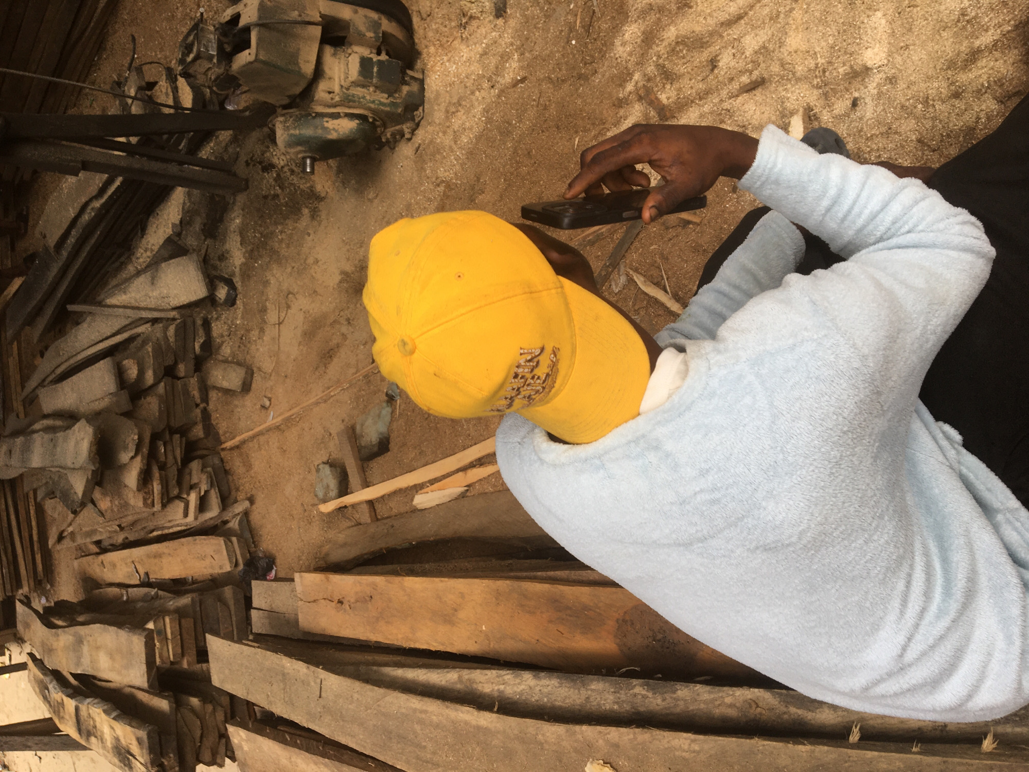 The image presents an overhead view of a person in a woodworking environment, likely a sawmill or carpentry workshop, located in Ado, Nigeria. The ground is predominantly covered in sand, dirt, and a generous layer of sawdust, mixed with numerous wood scraps and offcuts.
The primary subject, an individual with dark skin, is seen from behind and slightly to the side, wearing a bright yellow baseball cap that clearly displays the text "NEW YORK" on its front. They are dressed in a light blue, fuzzy long-sleeved shirt and dark pants. The person is holding a black smartphone with both hands, actively engaging with the device using their right index finger, suggesting they are texting, browsing, or making a call.
Surrounding the individual are various pieces of timber and wood fragments of different sizes, indicating a busy workspace. In the upper left portion of the frame, a dusty, green industrial machine with belts and gears is visible, characteristic of heavy-duty woodworking equipment like a sawmill or planer. The lighting suggests it is daytime. The overall scene depicts a moment of respite or personal activity within a laborious setting.